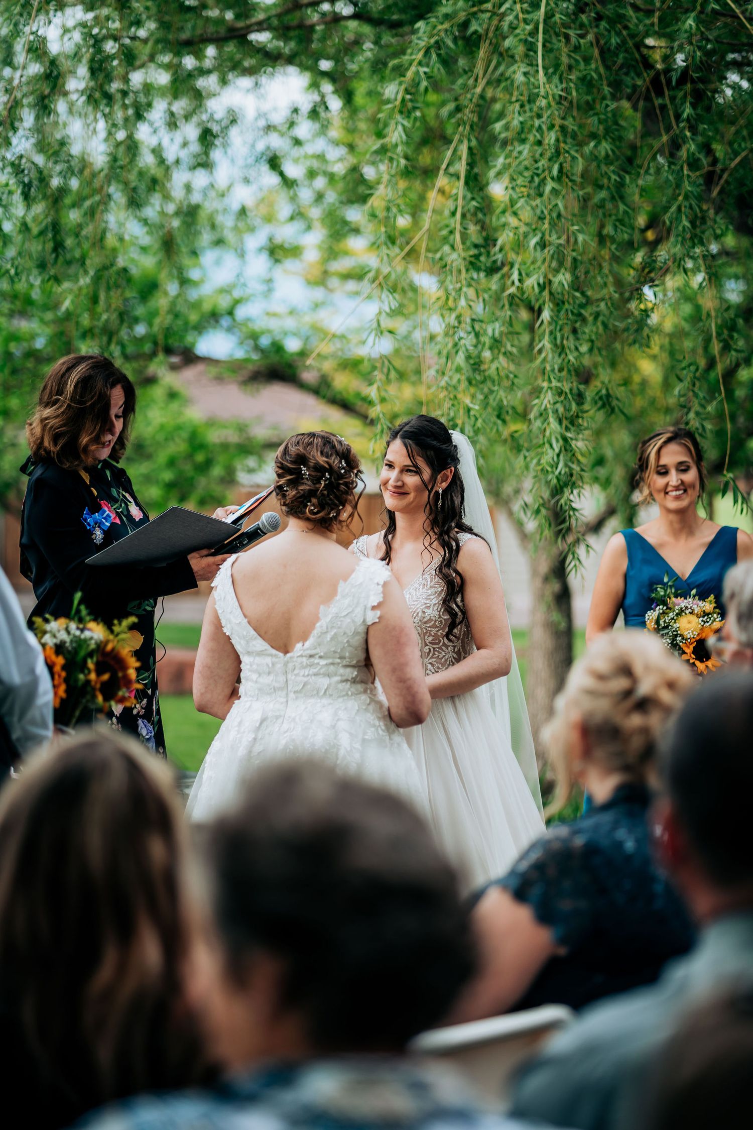 Outdoor wedding ceremony under lush green trees with guests watching the couple exchange vows.