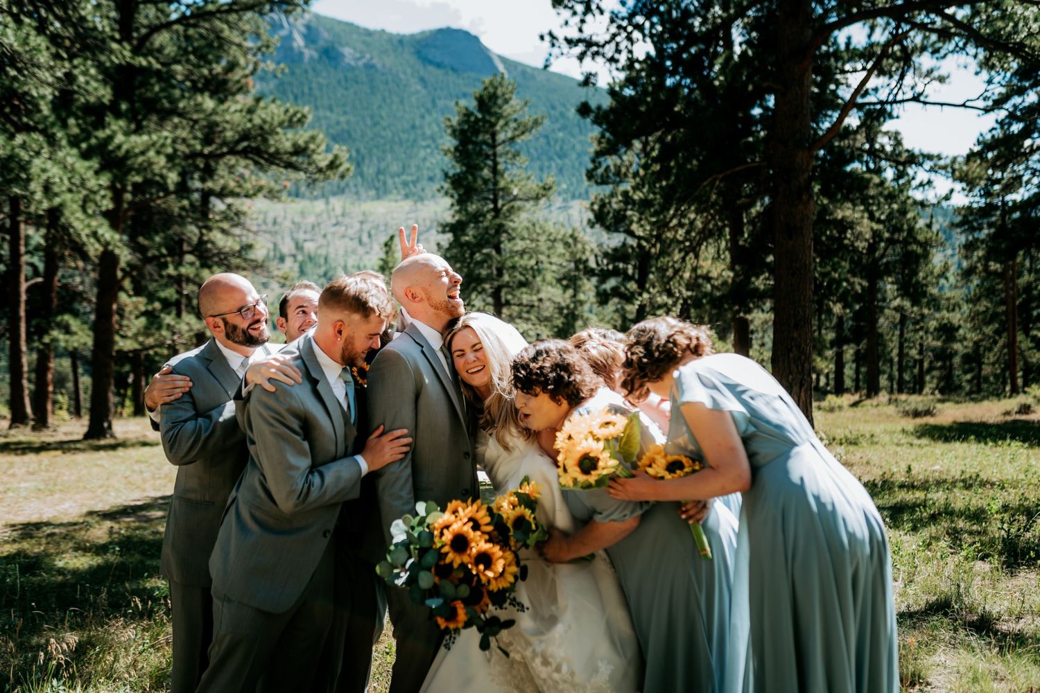 Wedding party gathers in a mountain meadow surrounded by evergreen trees.
