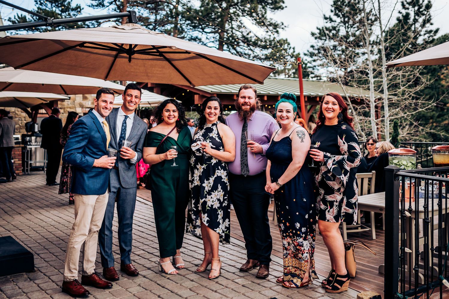 A group of wedding guests in formal attire pose together on a wooden deck during sunset.
