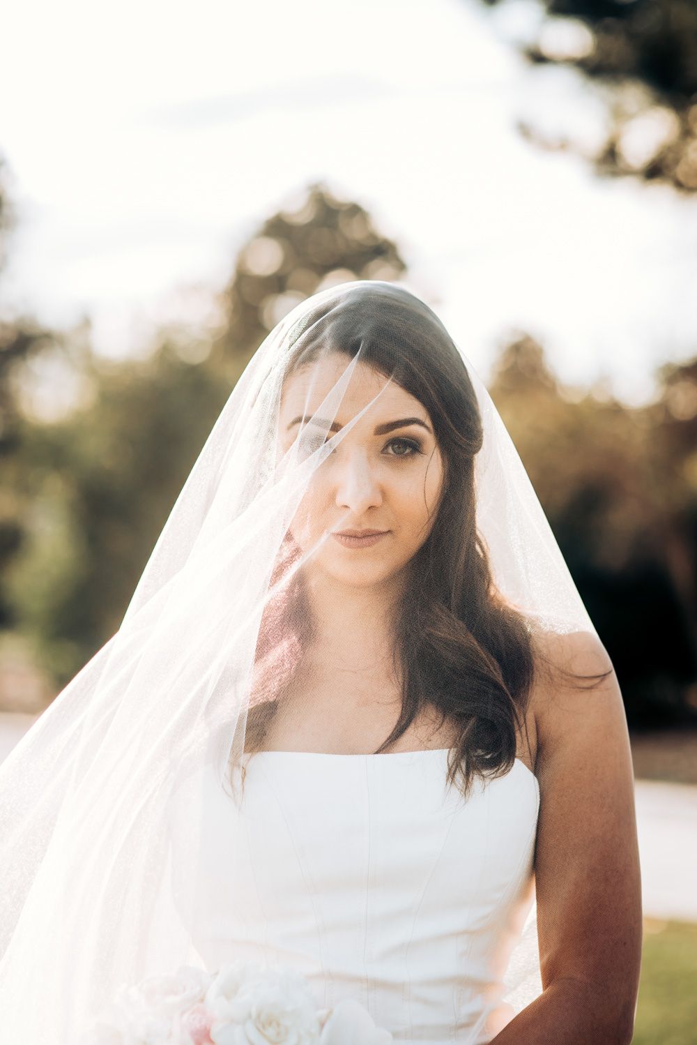 Elegant bride in white dress and long veil poses outdoors in natural lighting.
