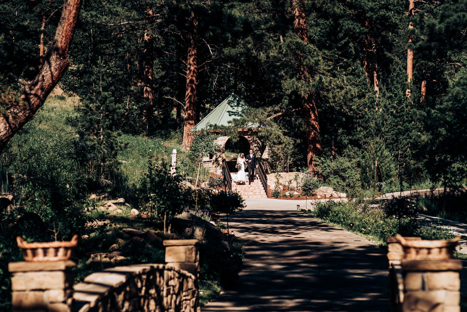 A scenic stone bridge pathway through pine trees leads to a rustic outdoor ceremony space.