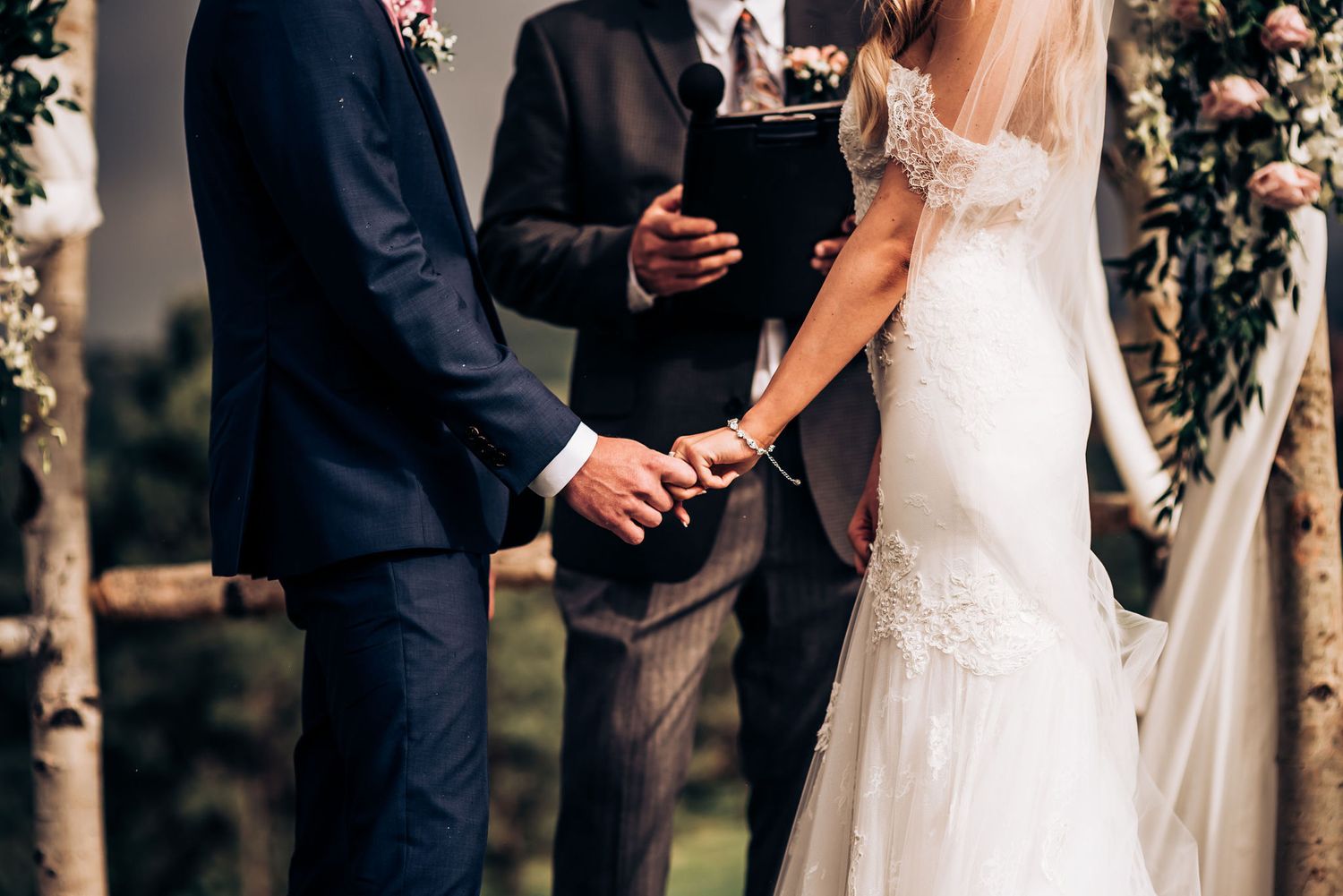 Close-up of couple holding hands during wedding ceremony with officiant in background.