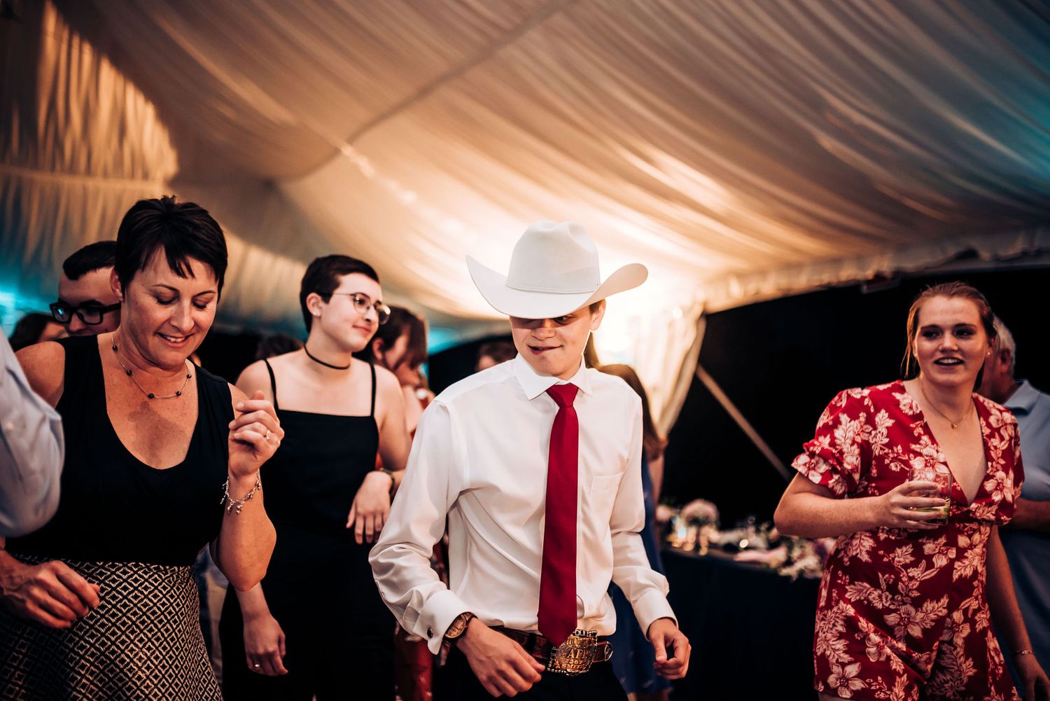 Wedding guests dancing under a draped tent with atmospheric lighting at an evening reception.
