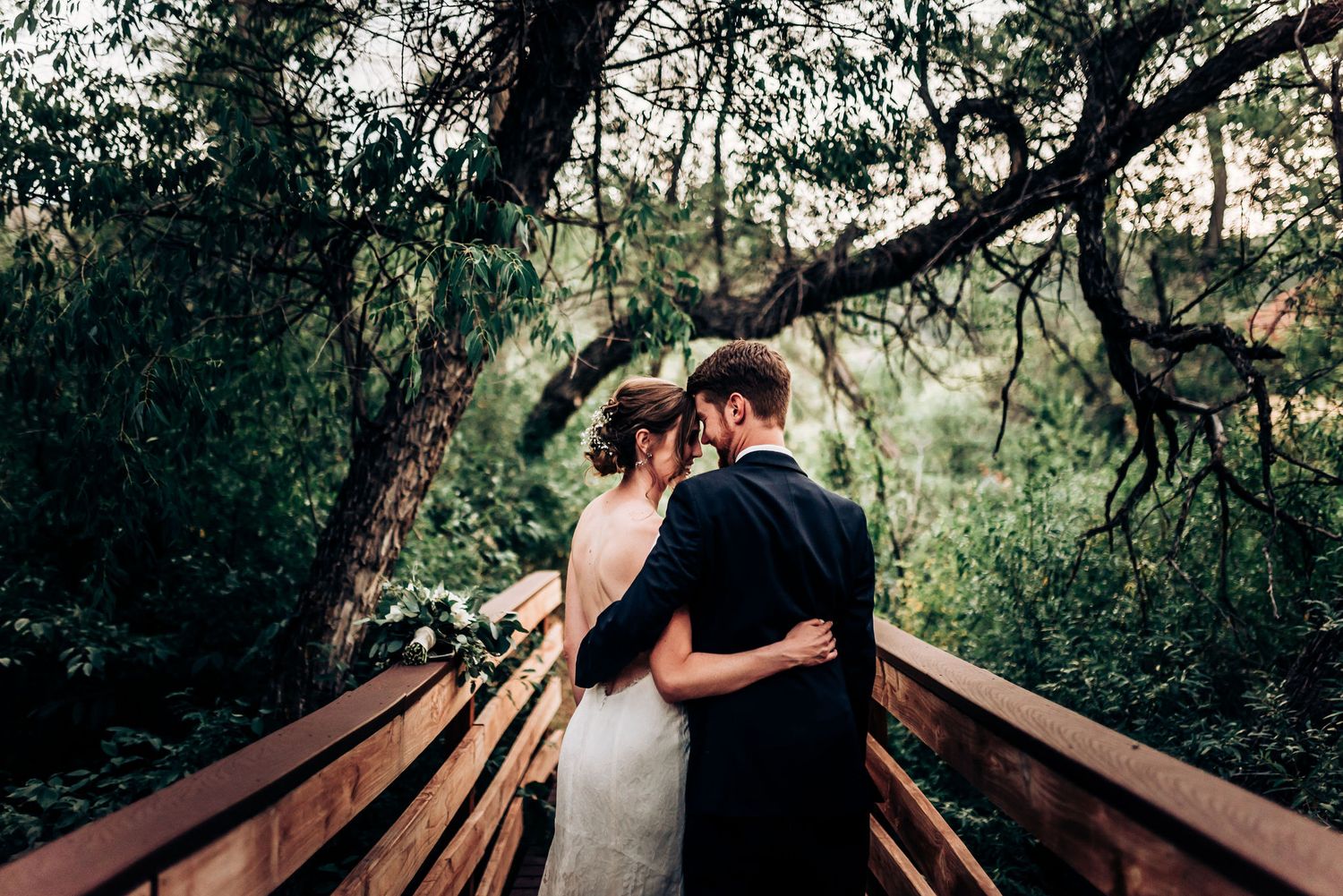 Romantic wedding couple embracing on wooden bridge under tree branches at sunset.