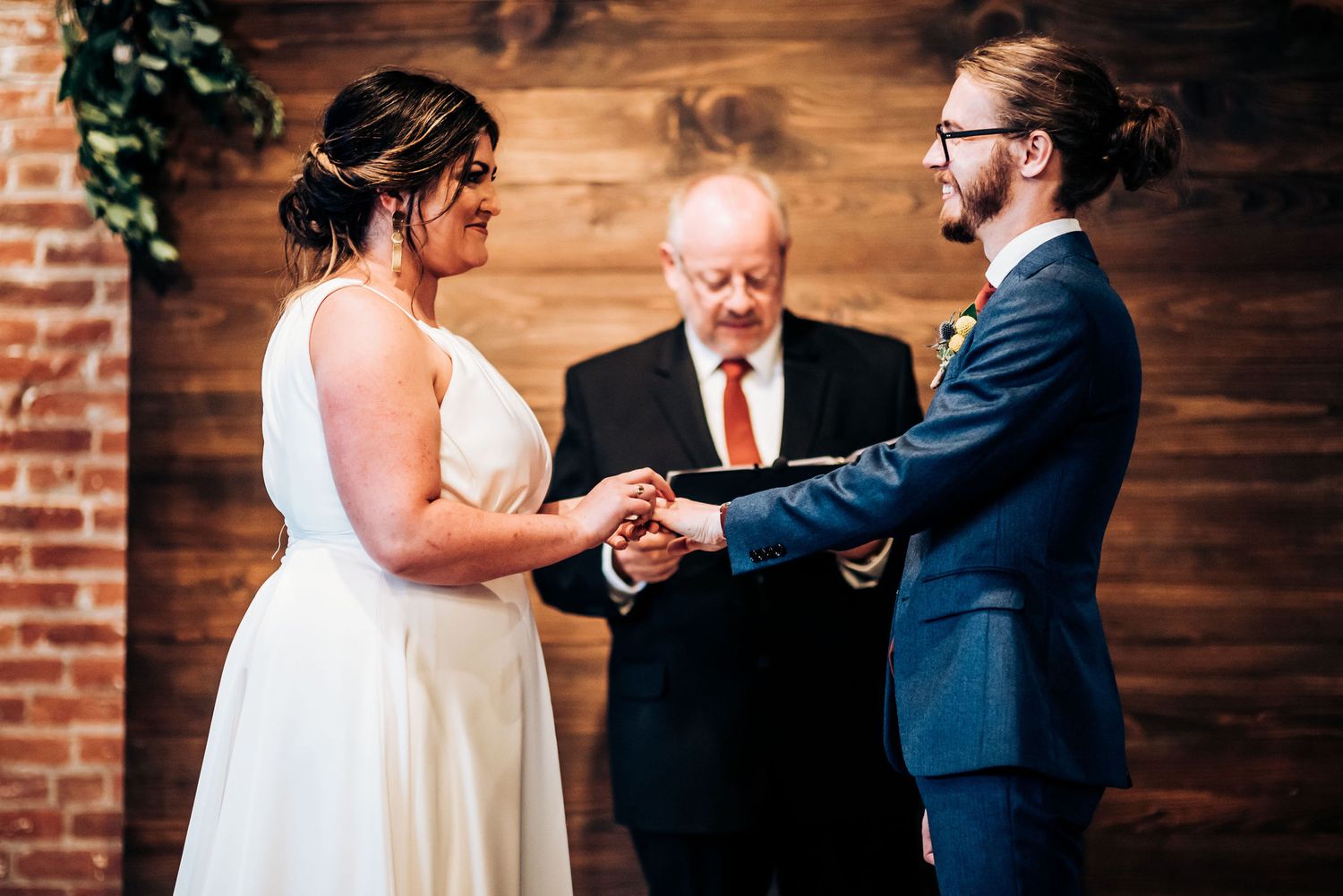 Intimate wedding ceremony moment as couple joins hands before officiant against wood paneled wall.
