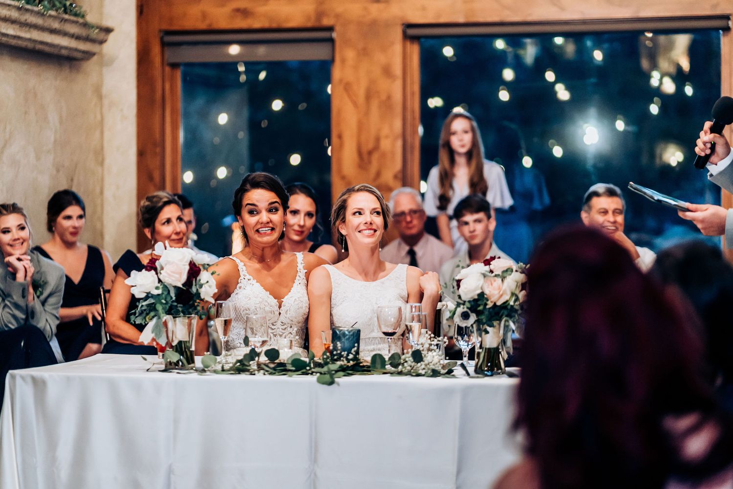 Wedding party members seated at head table with floral centerpieces and twinkling lights in background at reception.