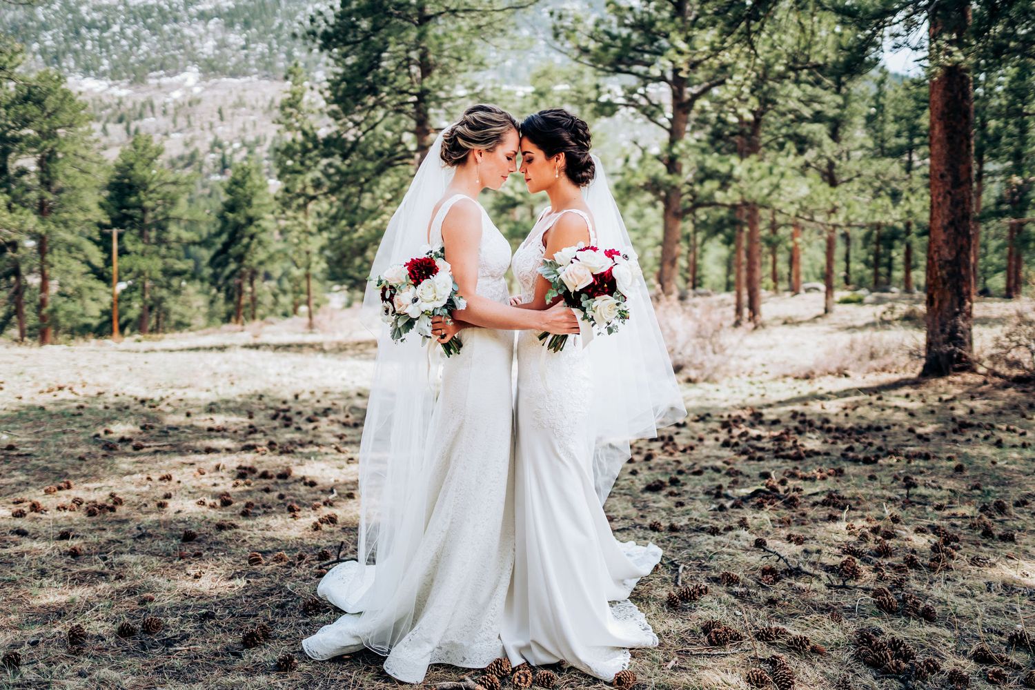 Newlyweds share tender moment in white dresses against mountain forest backdrop.