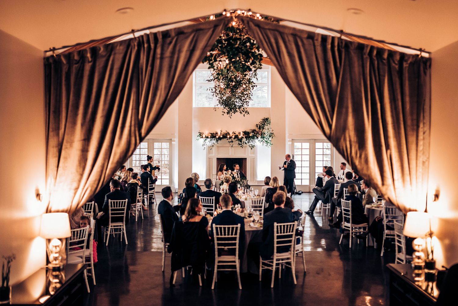 Elegant wedding reception venue with dramatic drapes, chandeliers and greenery decorations.