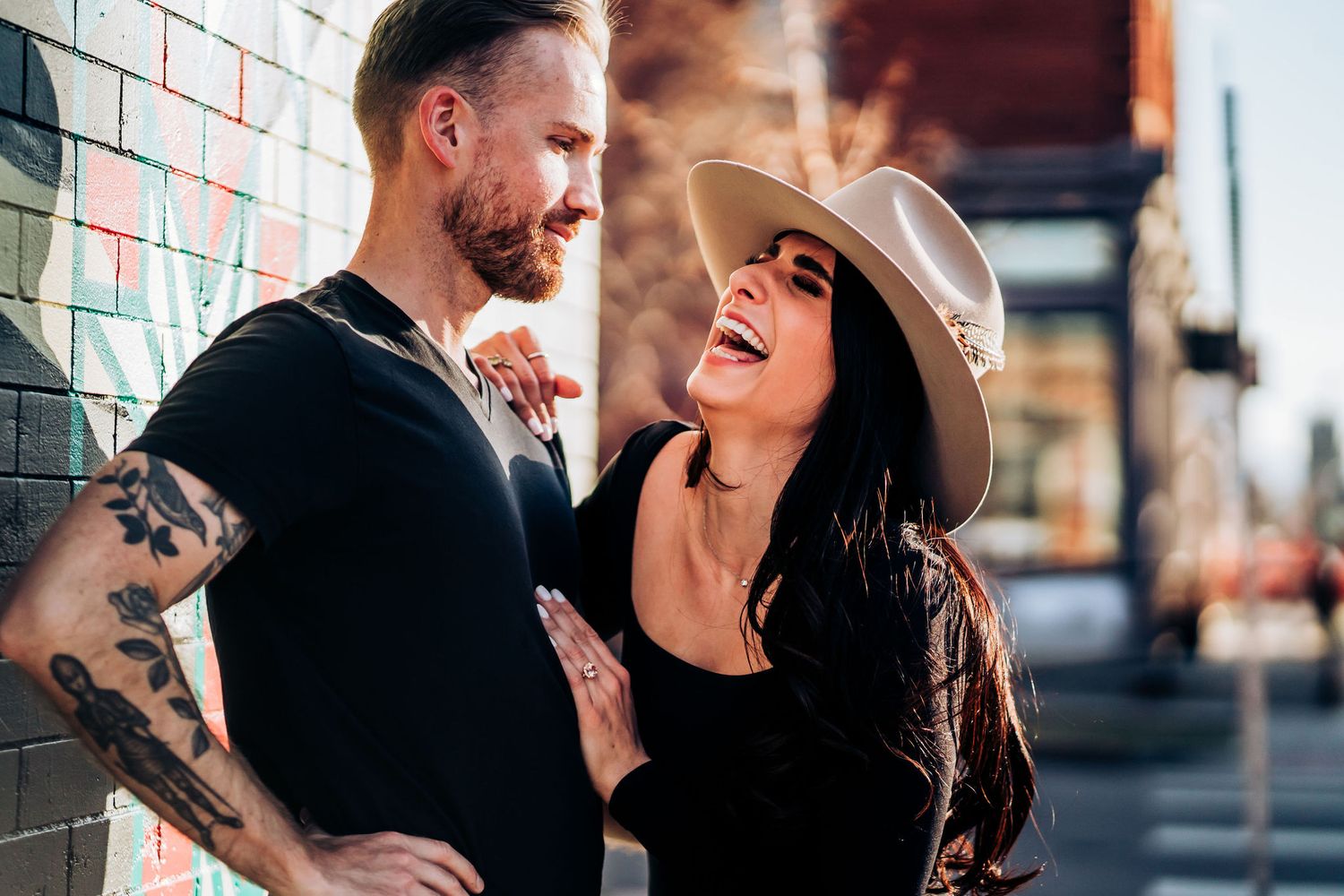 Couple shares laughing moment against brick wall in urban setting.