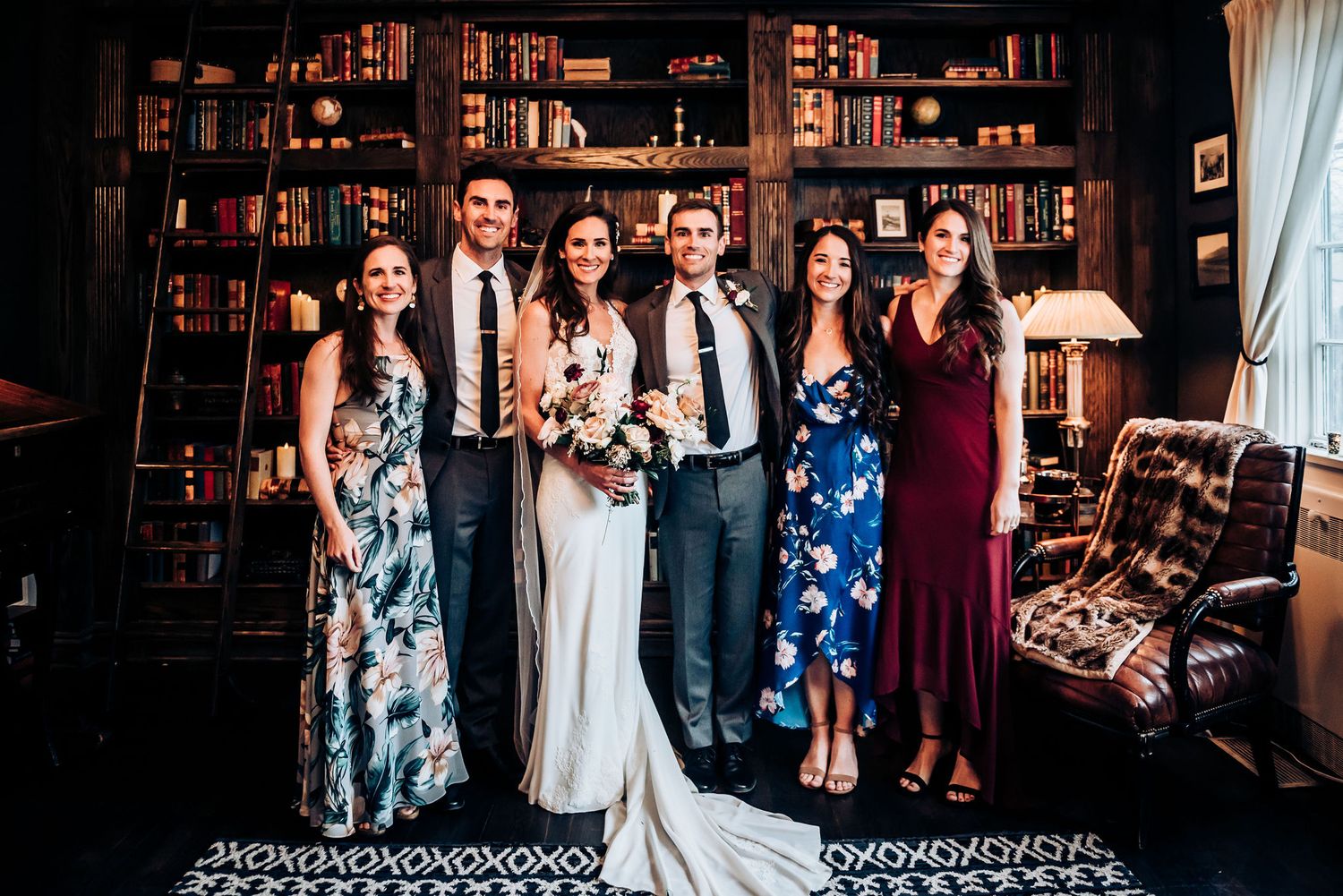 Wedding party poses in front of vintage library bookshelves wearing elegant floral dresses and formal attire.