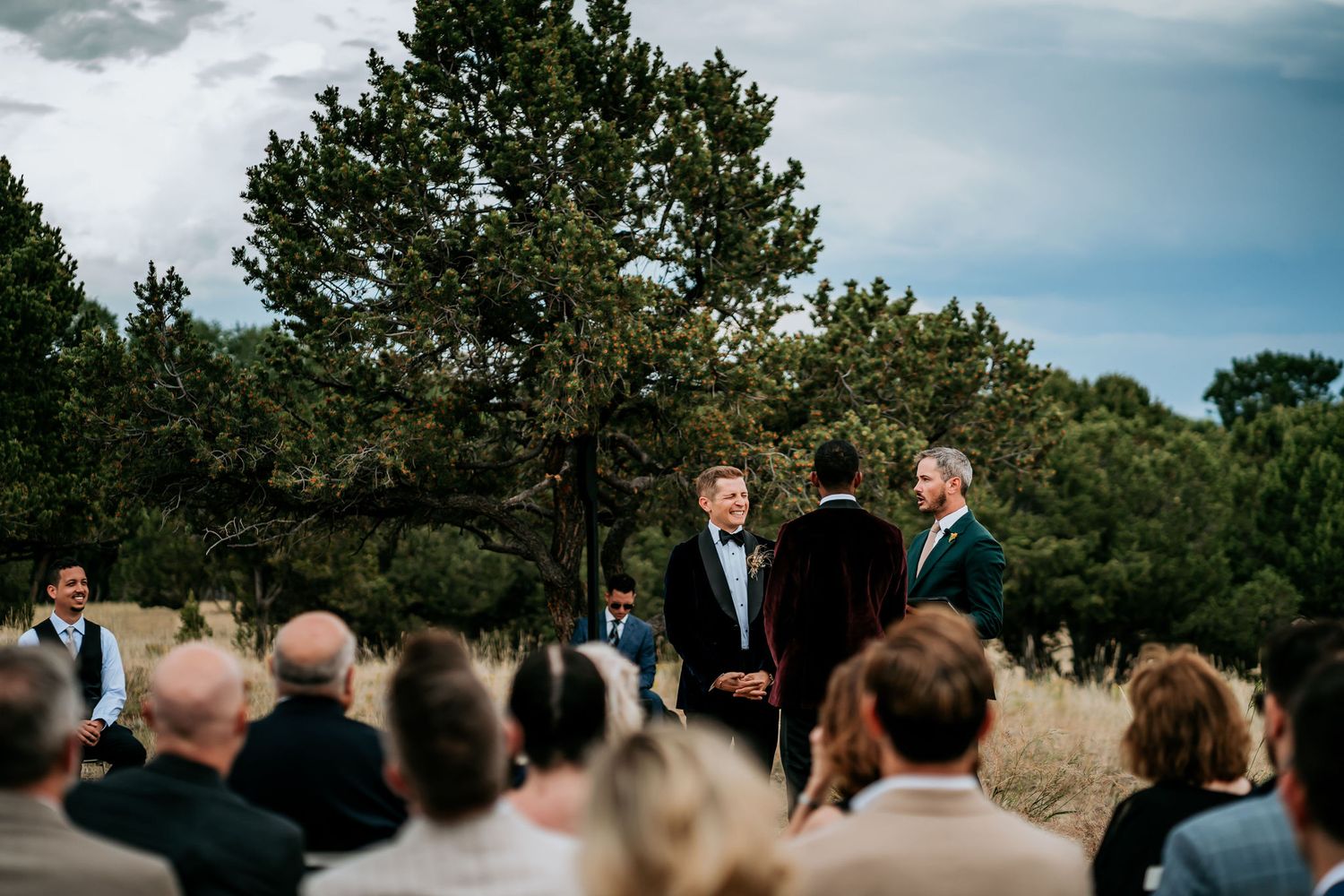 Wedding ceremony taking place outdoors with guests seated and trees in background.