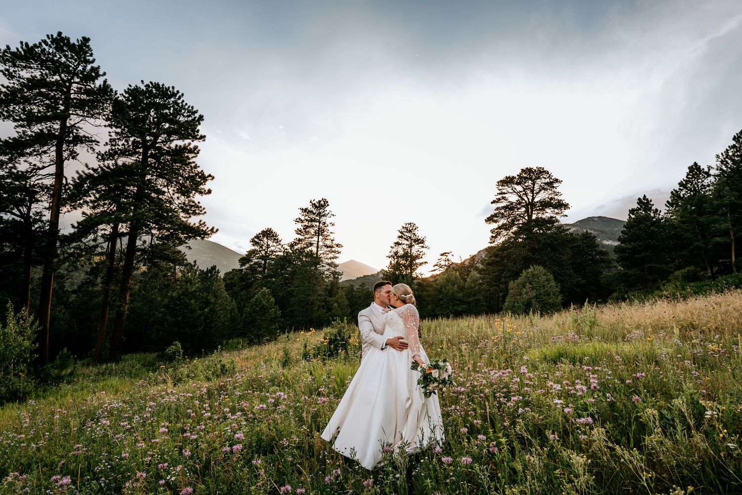 Romantic sunset wedding portrait in mountain meadow with evergreen trees silhouetted against sky.