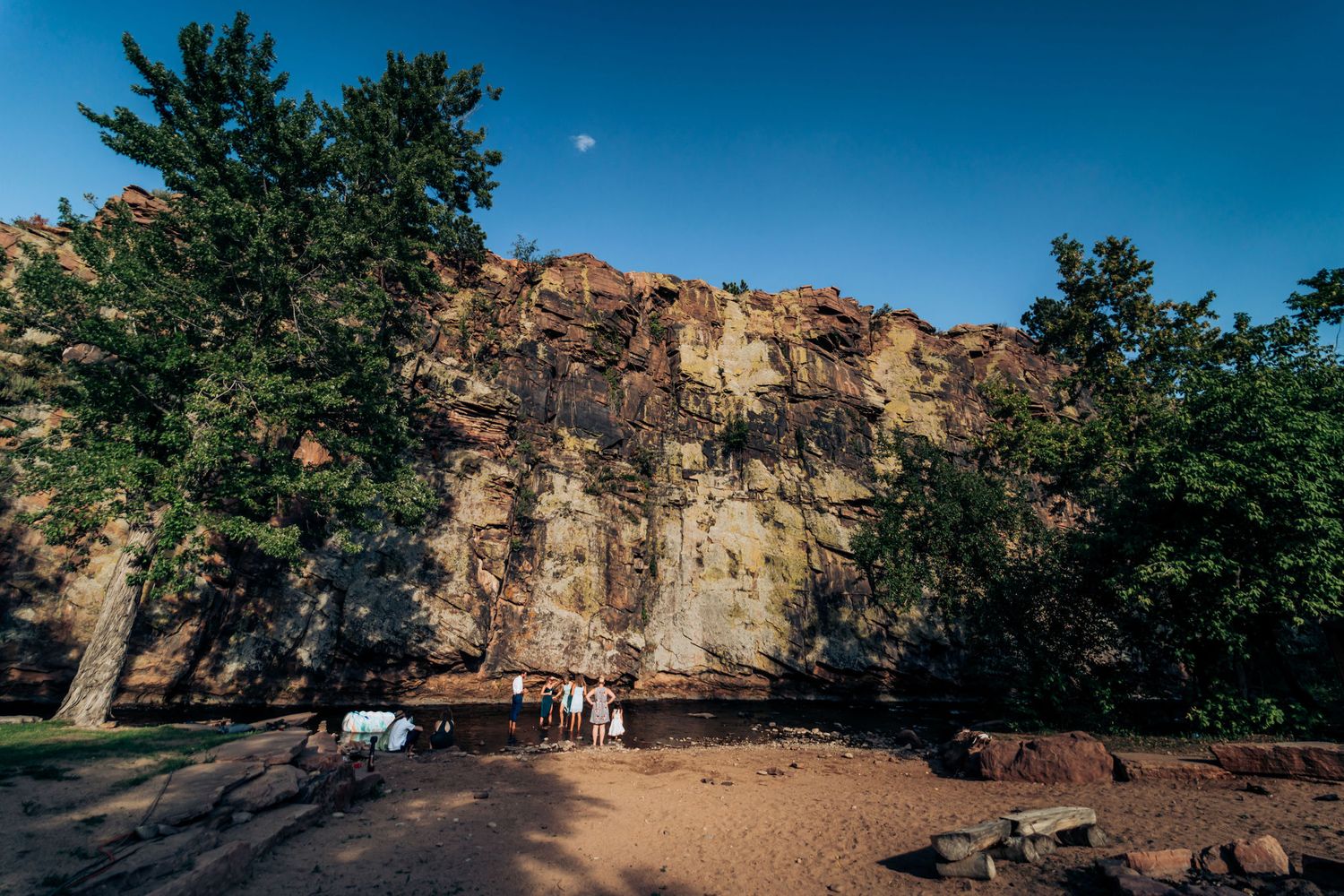 Majestic rocky cliff face towers over a park area with pine trees under bright blue sky.