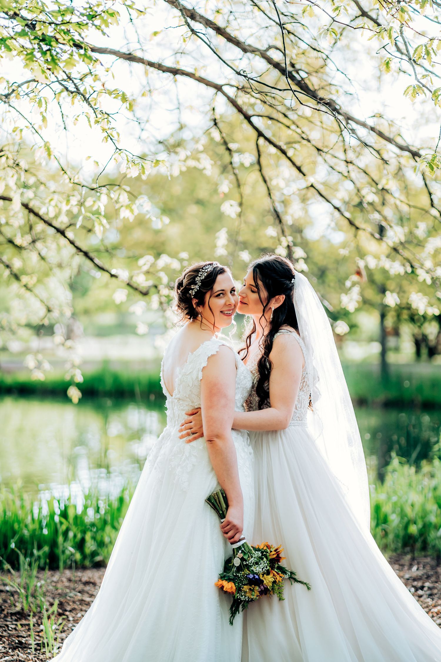 Wedding couple in white gowns embraces under flowering spring tree branches near water.
