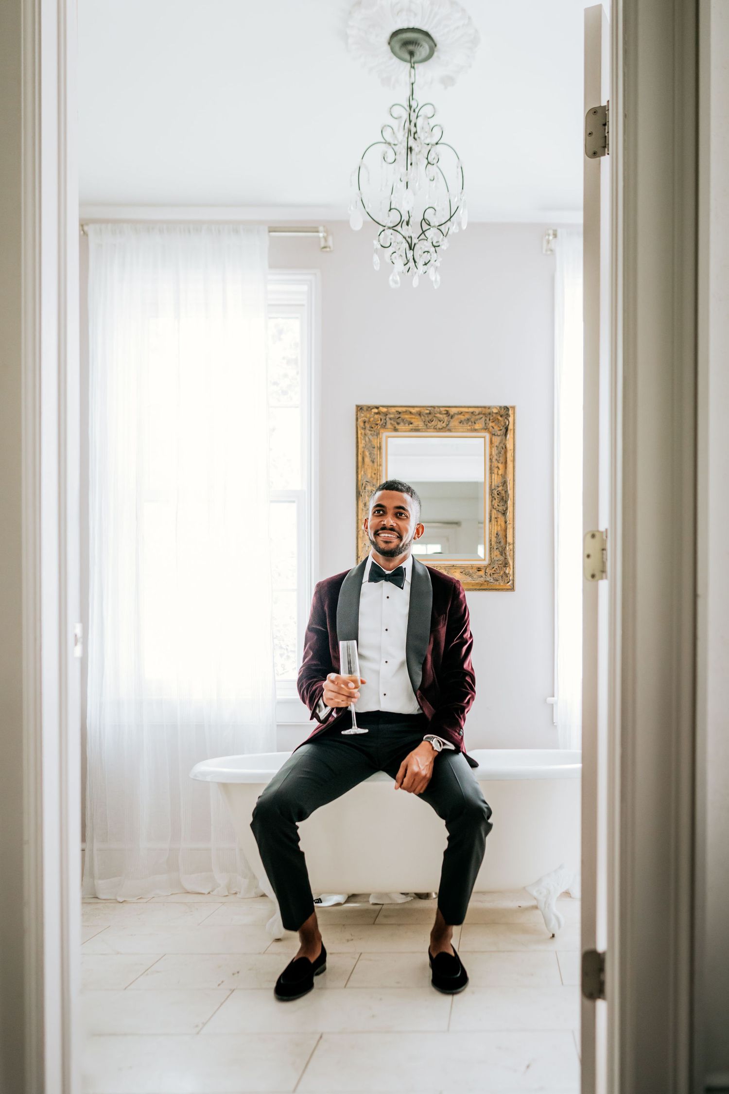 A person in formal attire sits on a ledge in an elegant white room with a chandelier.