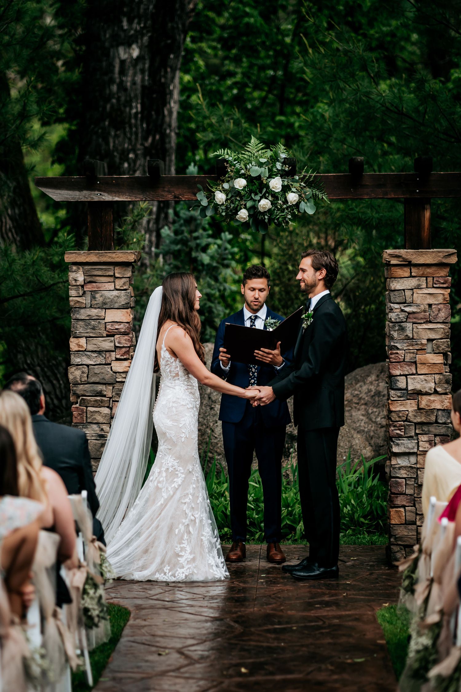 An outdoor wedding ceremony takes place between stone pillars with white floral decorations.