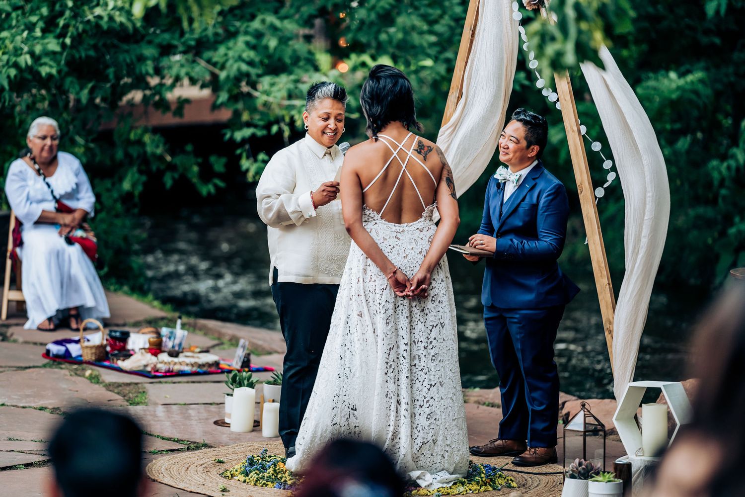 An intimate outdoor wedding ceremony unfolds beneath a rustic wooden arch with flowing white fabric draped on sides.