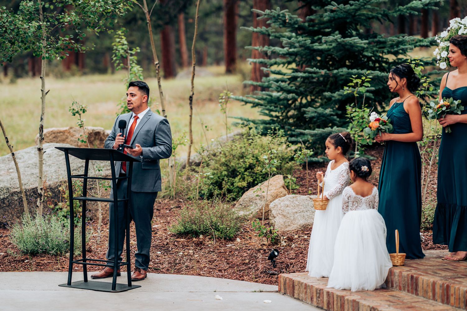 A wedding speech being delivered at an outdoor ceremony surrounded by evergreen trees.