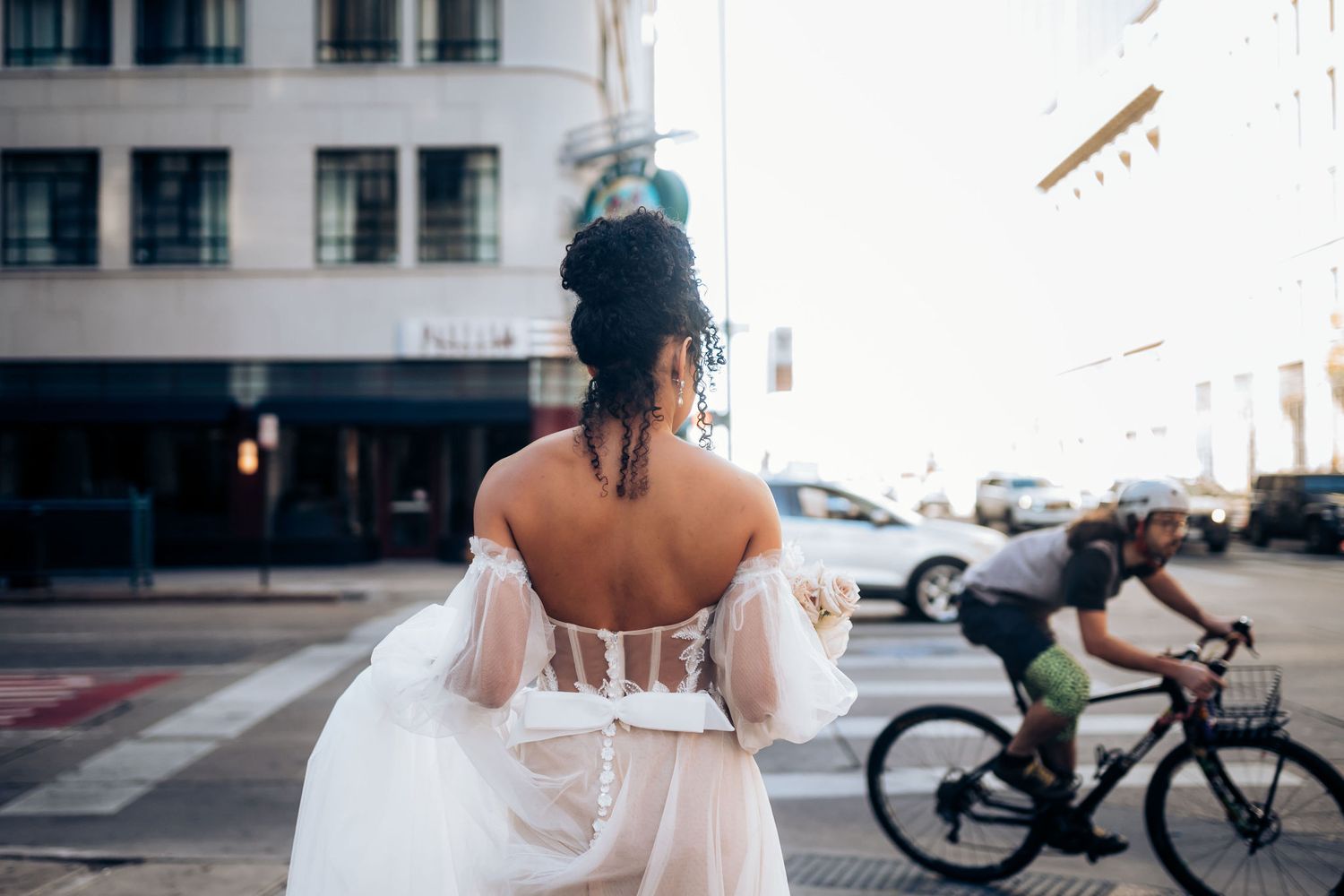 Back view of bride in white dress with sheer details as cyclist passes on urban street.