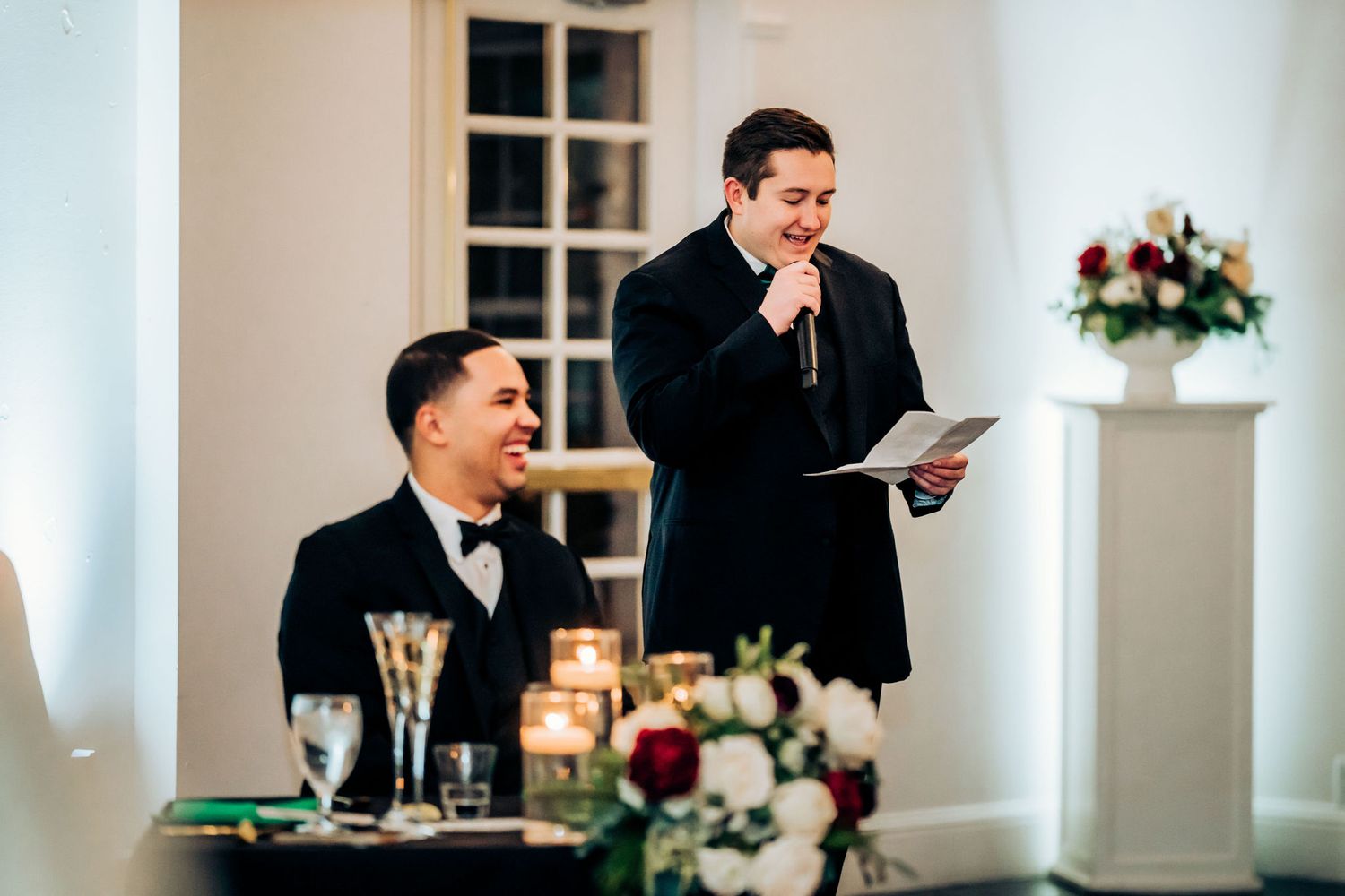 Two people in formal attire during wedding reception speeches near floral centerpieces.