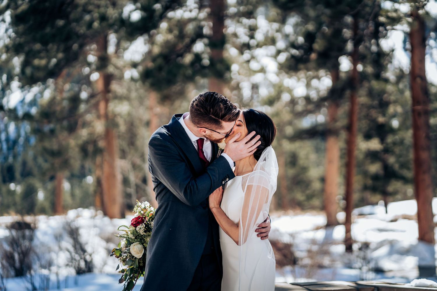 An intimate wedding moment captured in a snowy forest setting with pine trees in the background.