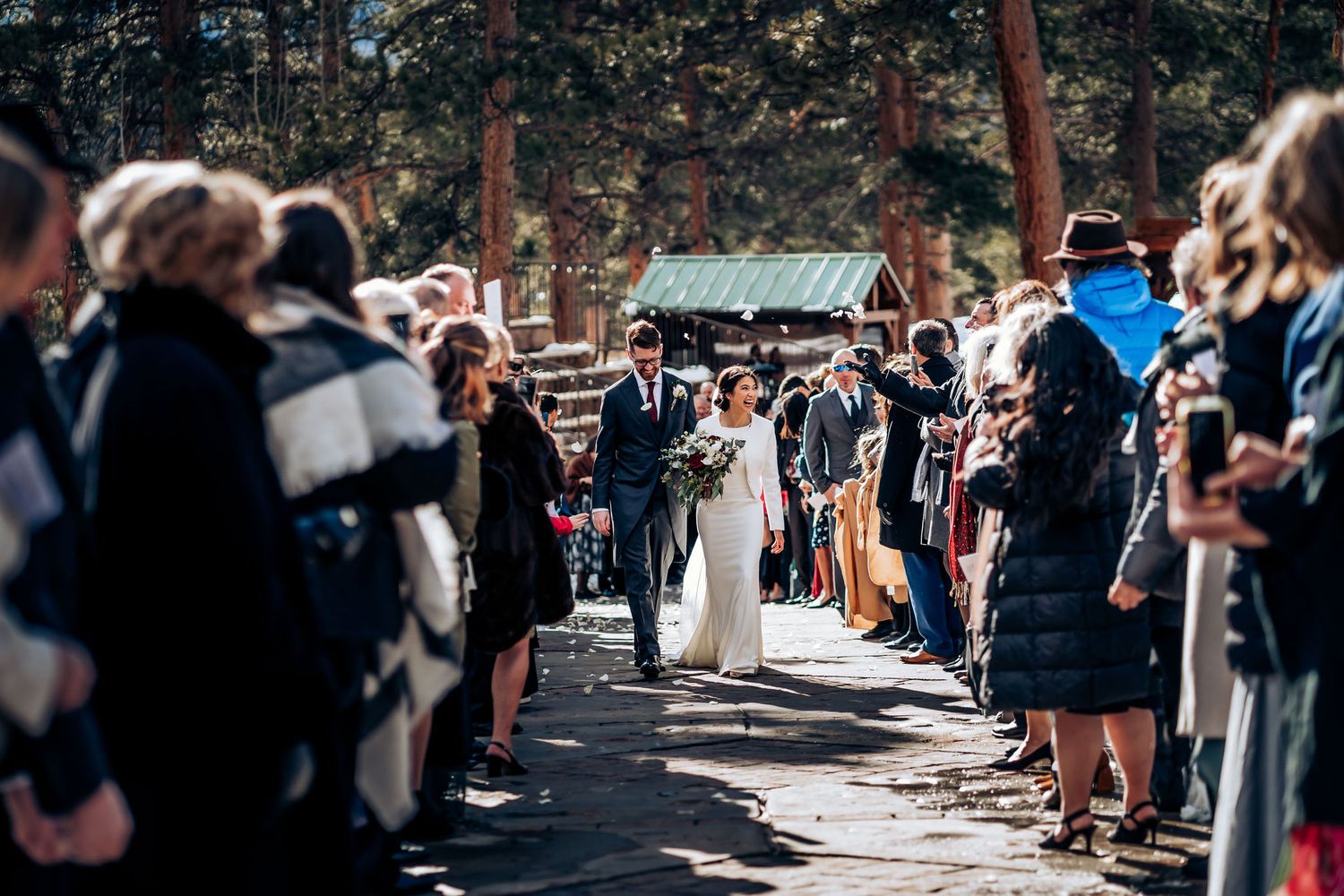 Wedding party processes through crowd-lined path with rustic cabin visible in background.