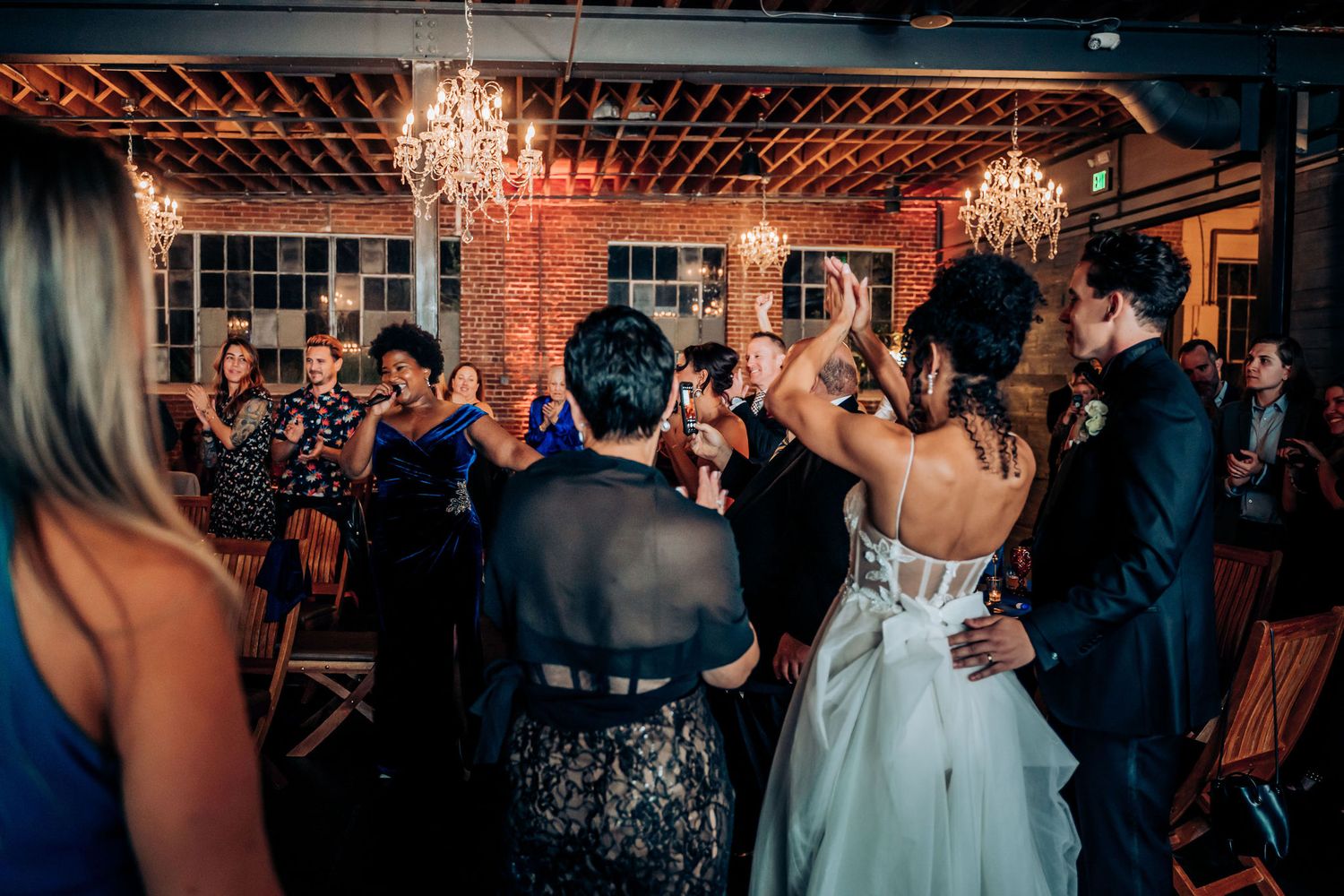 Wedding guests dance and celebrate under crystal chandeliers in a rustic brick venue.