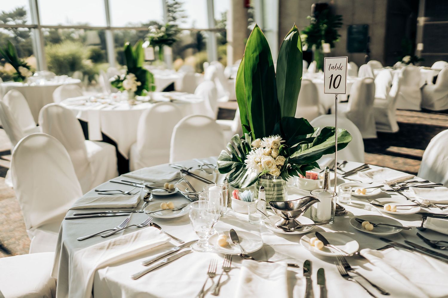 A formal place setting with white linens, elegant tableware, and tropical leaf centerpiece.