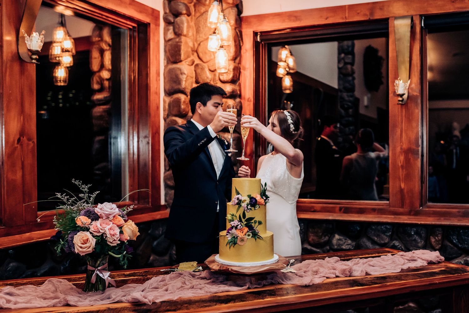Couple tasting their wedding cake during reception in a rustic lodge setting with warm lighting.