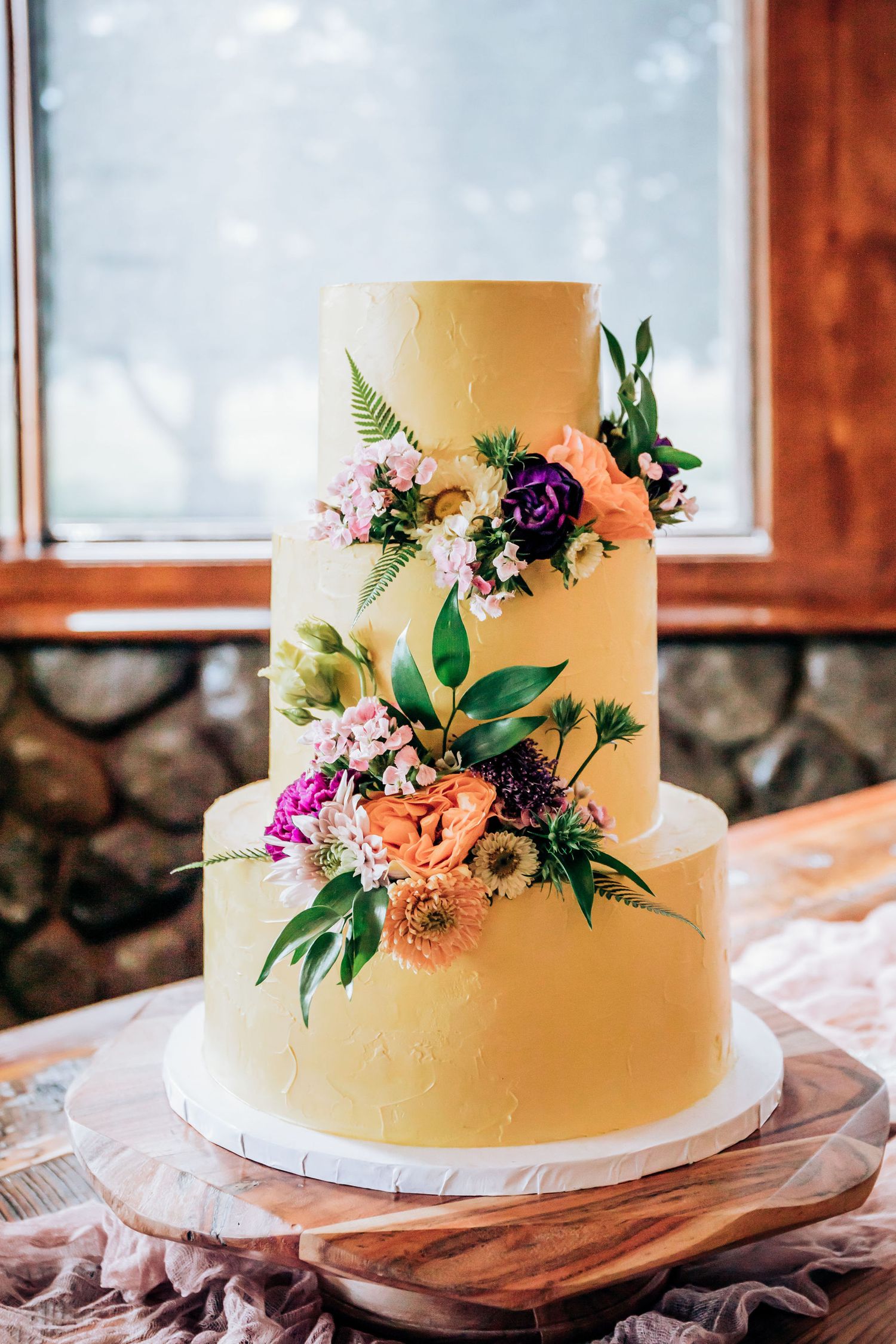 Elegant three-tier wedding cake decorated with fresh flowers and greenery on wooden stand.