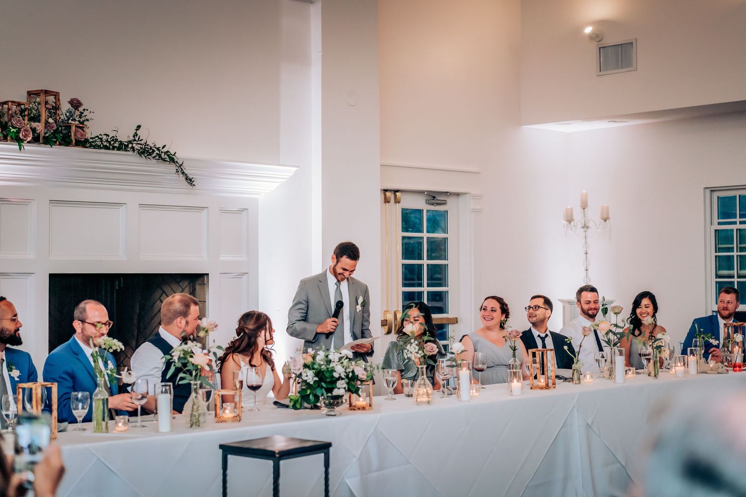 Wedding reception guests enjoy dinner and speeches at a white-decorated banquet table with floral centerpieces and candles.