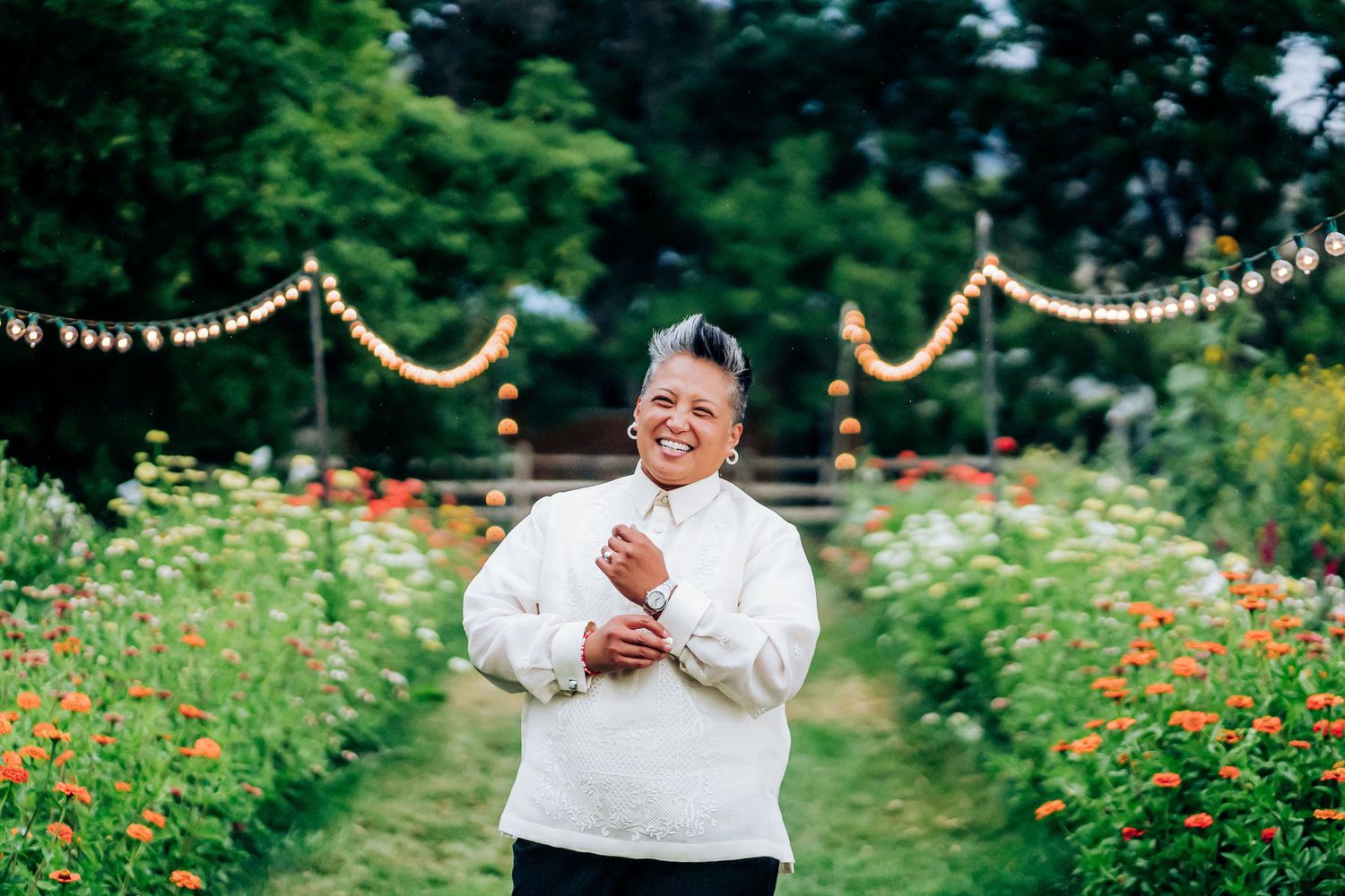 Person in white sweater laughing joyfully in a garden path decorated with string lights.