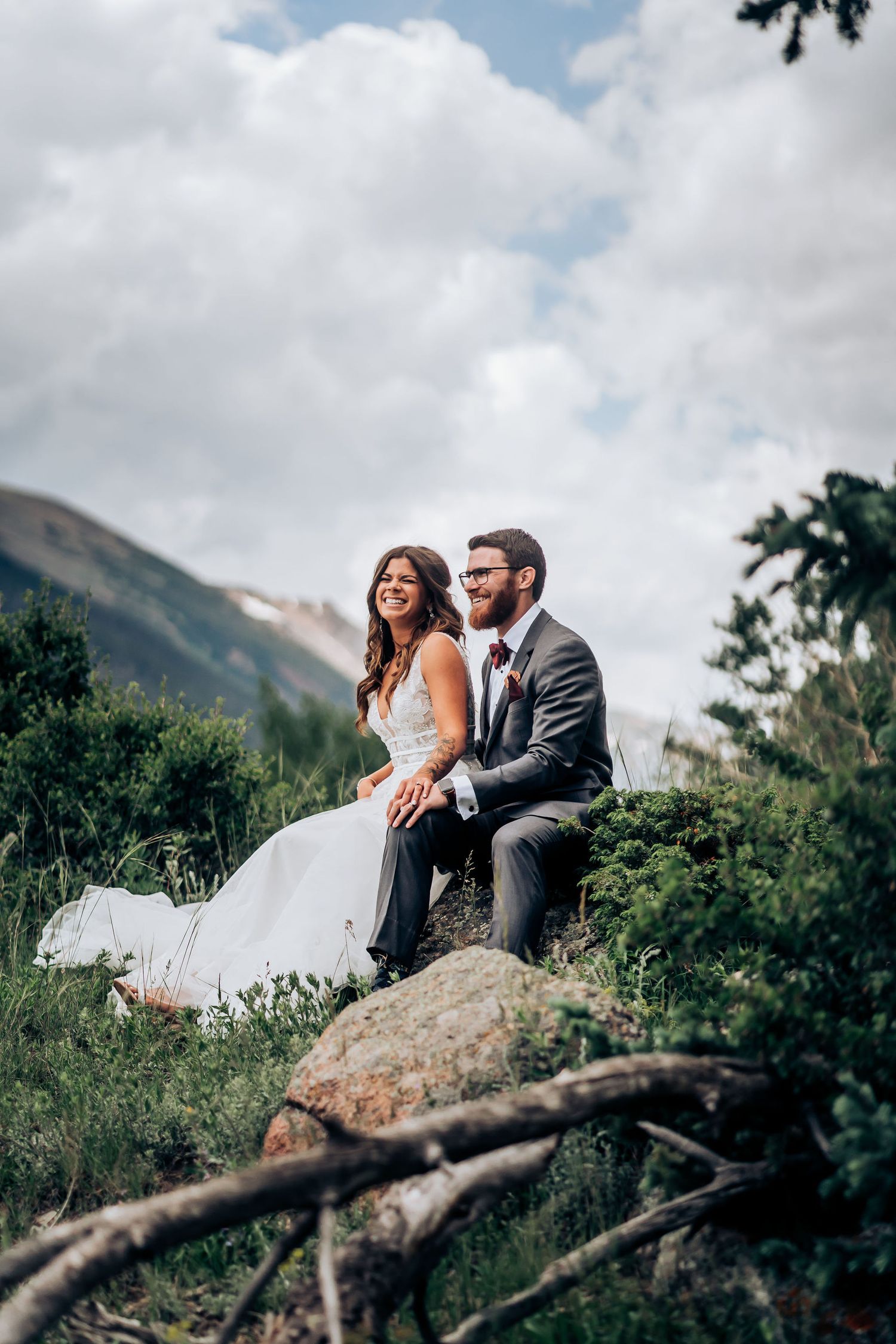 A wedding couple perches on a rock against a backdrop of majestic mountain peaks and cloudy skies.