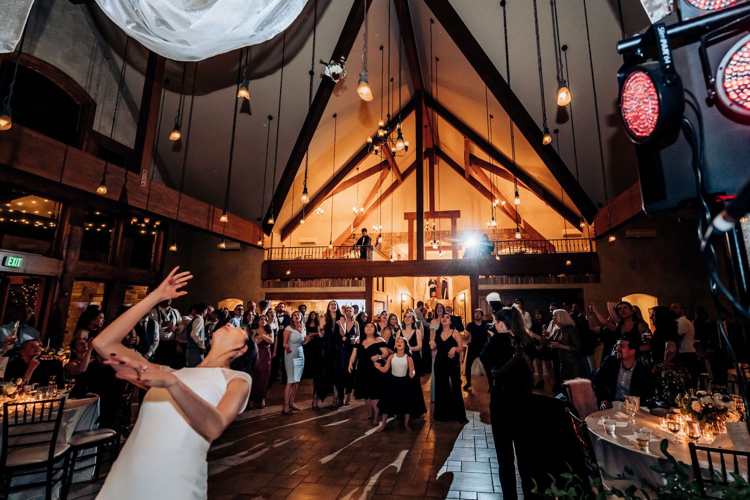 Wedding celebration with guests dancing under soaring wood beam ceilings and string lights.