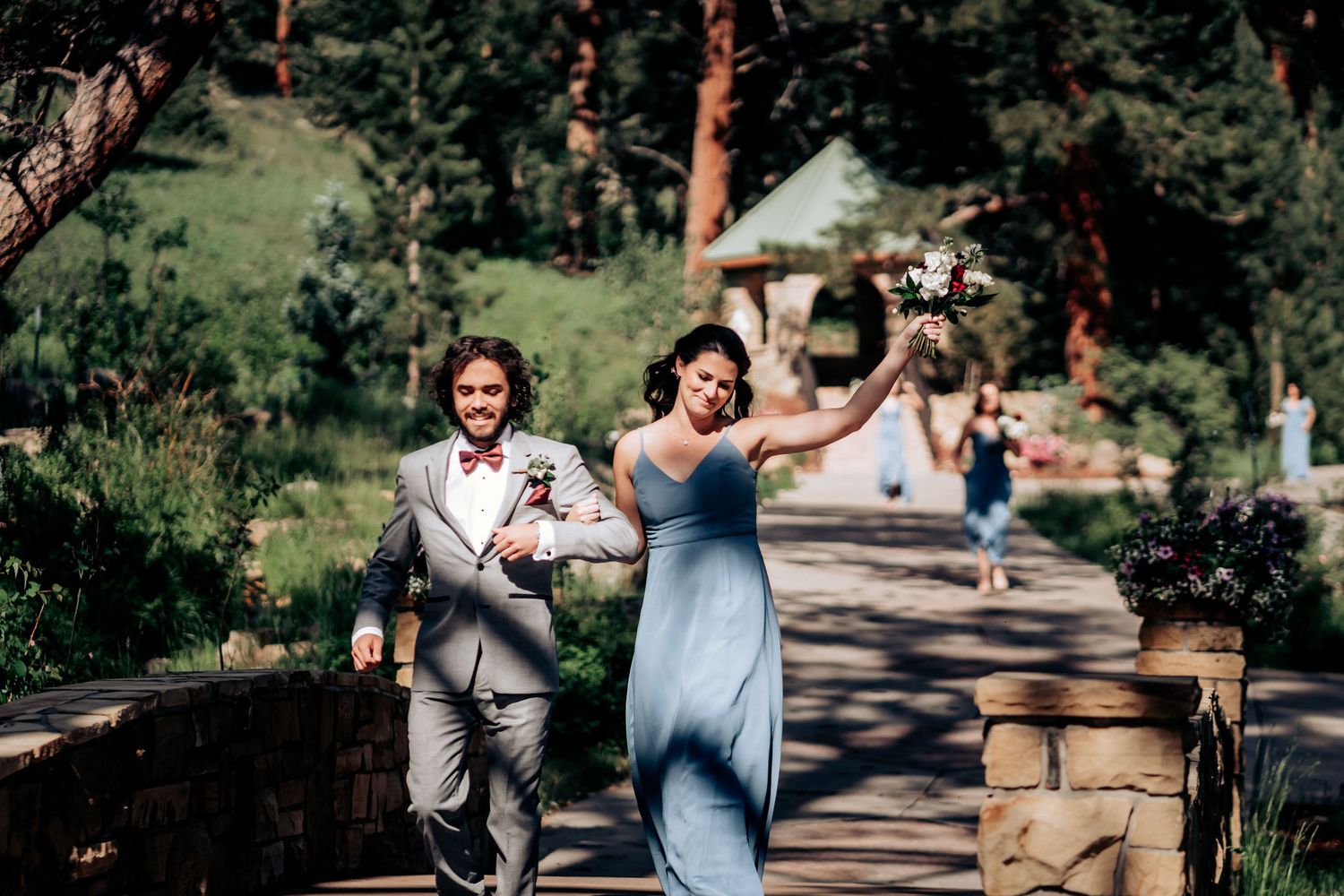 Wedding party members running joyfully down a stone path in a mountain setting.