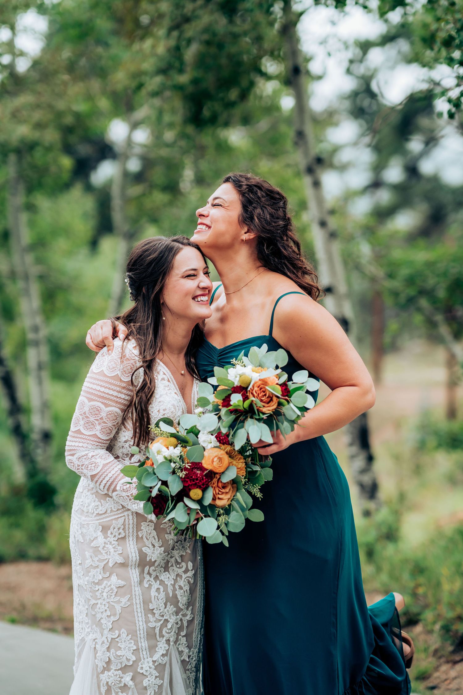 Two women share a joyful laugh while holding vibrant wedding bouquets outdoors.