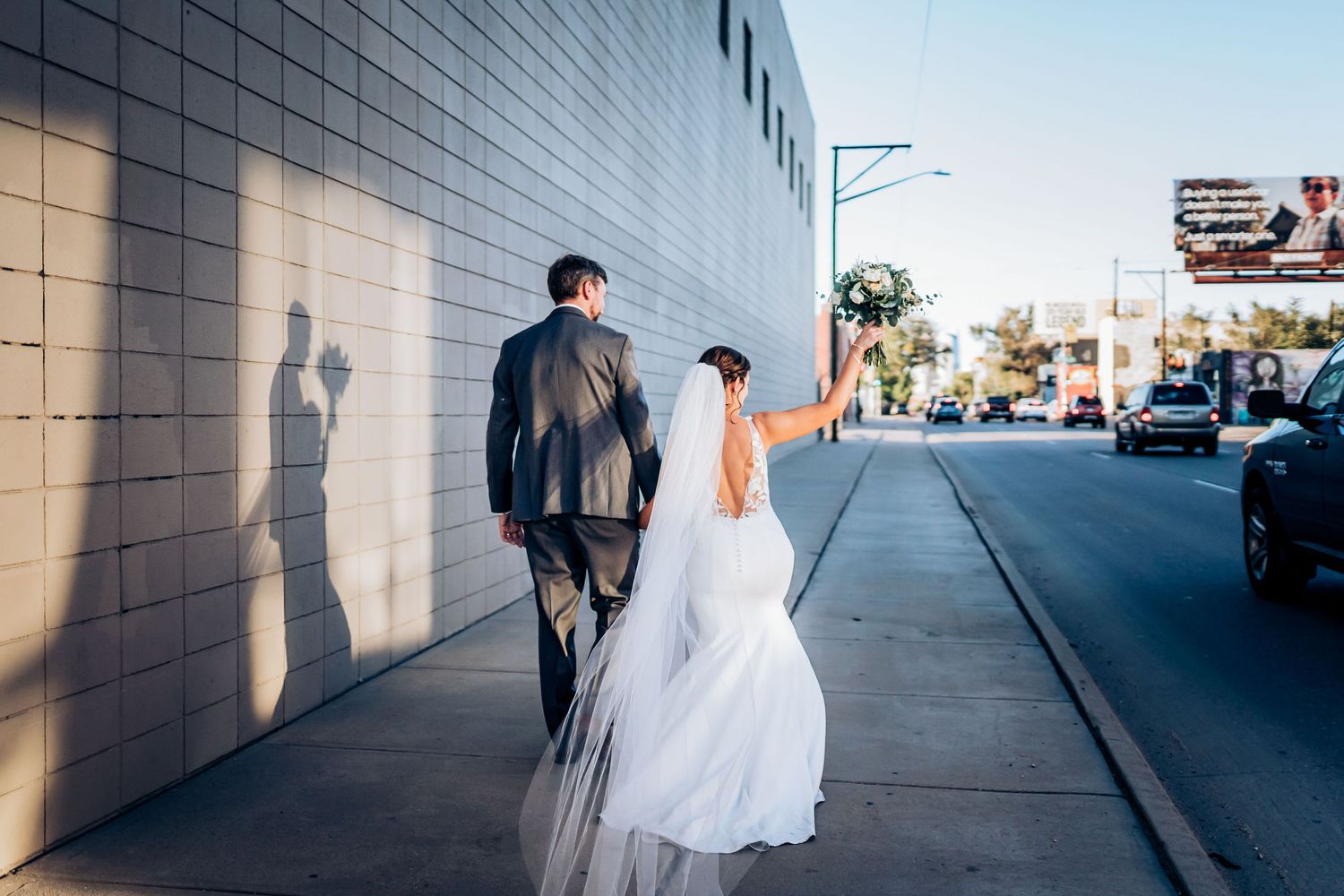 Wedding couple walking down urban sidewalk with dramatic shadows on white wall.