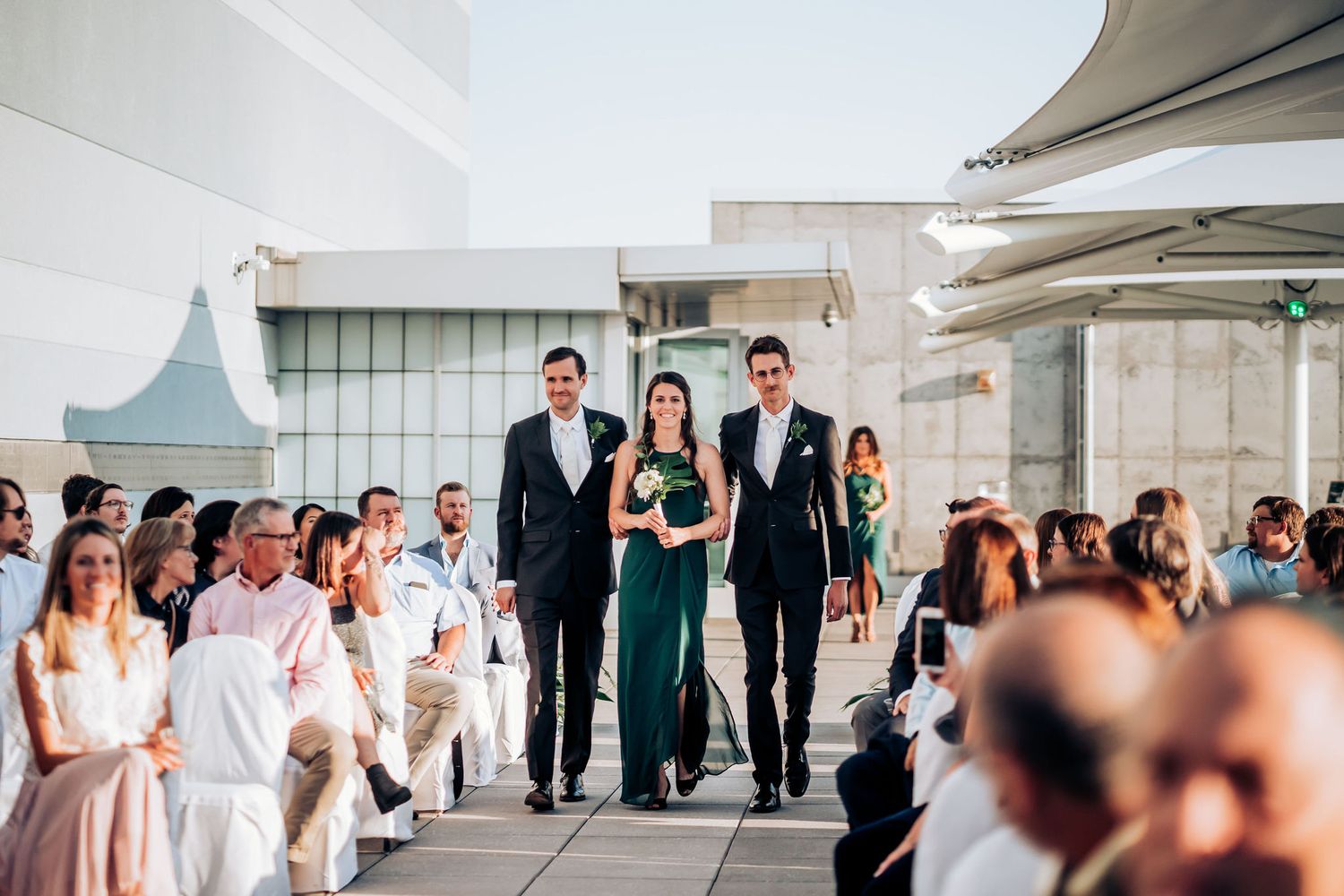 Wedding processional on a modern rooftop venue with seated guests in white and black attire.