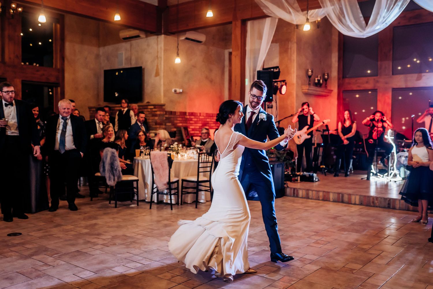 A couple performs an energetic dance move during their wedding reception as guests look on.