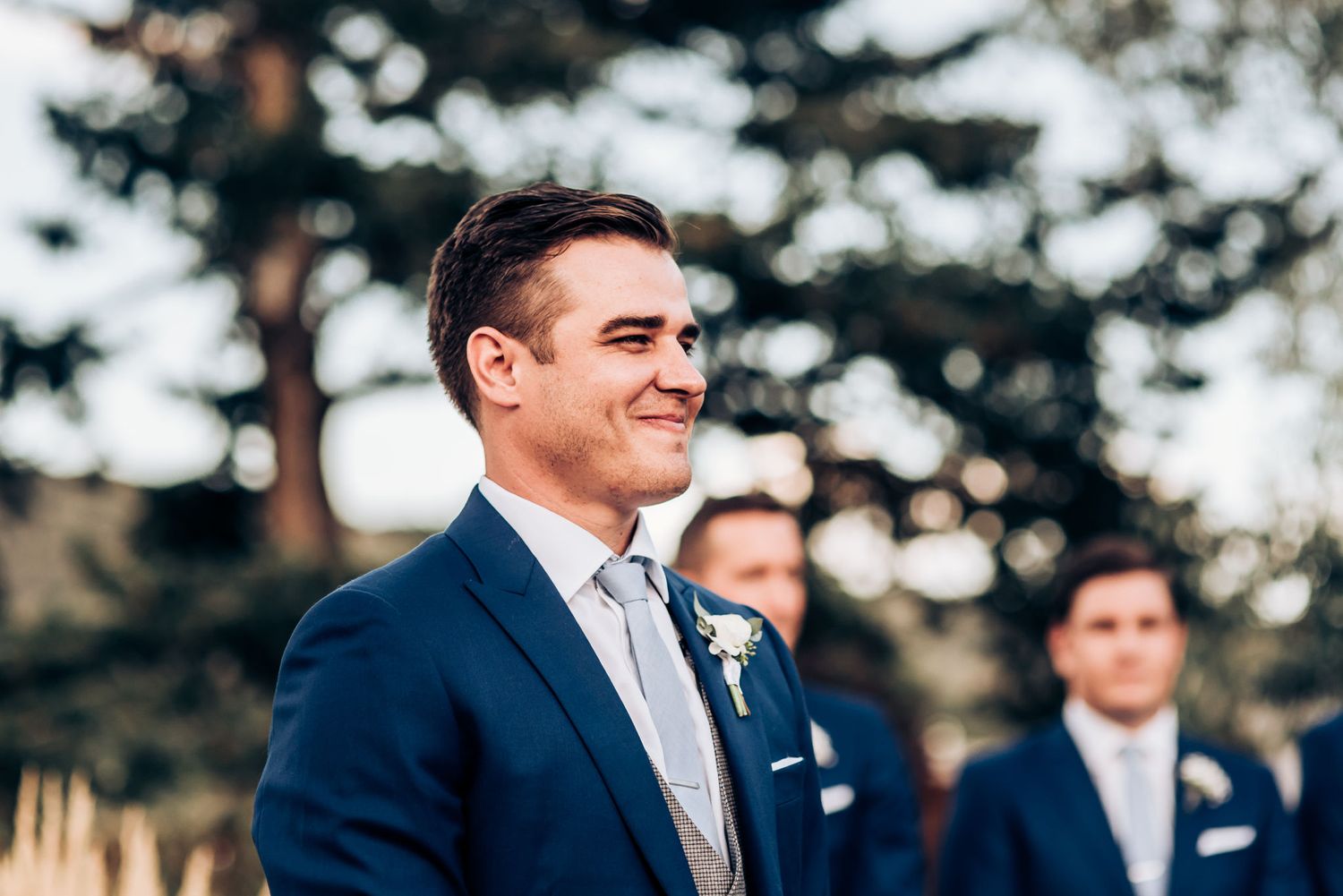Wedding participant in navy suit standing during outdoor ceremony with greenery in background.