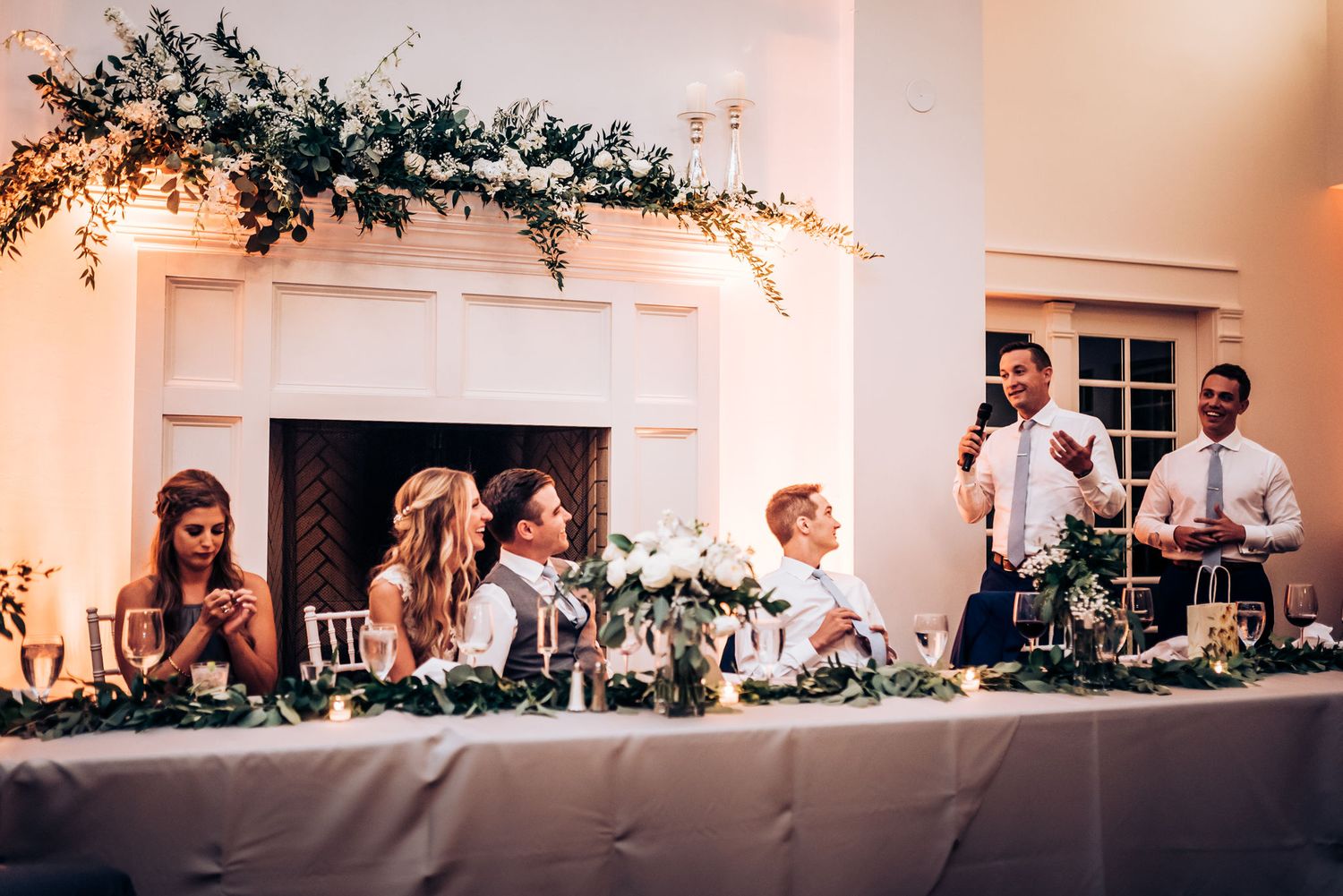 Wedding party seated at decorated head table during reception with greenery and warm lighting.