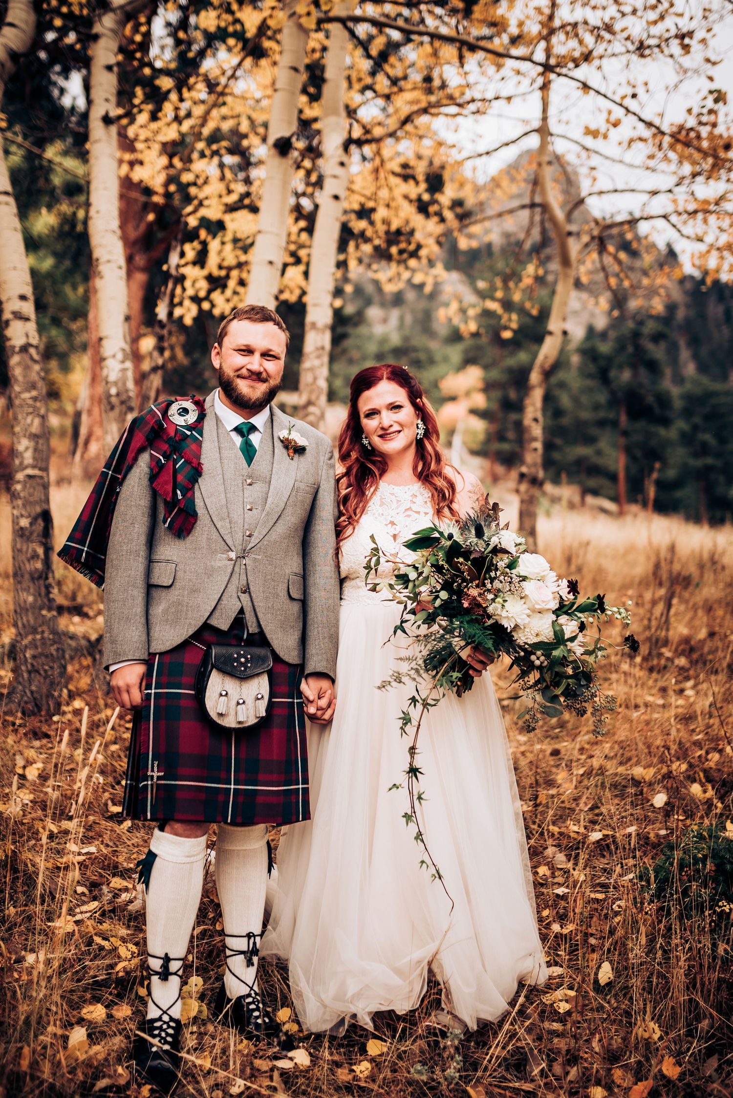A traditional Scottish wedding couple poses in autumn forest with groom in kilt and bride holding floral bouquet.