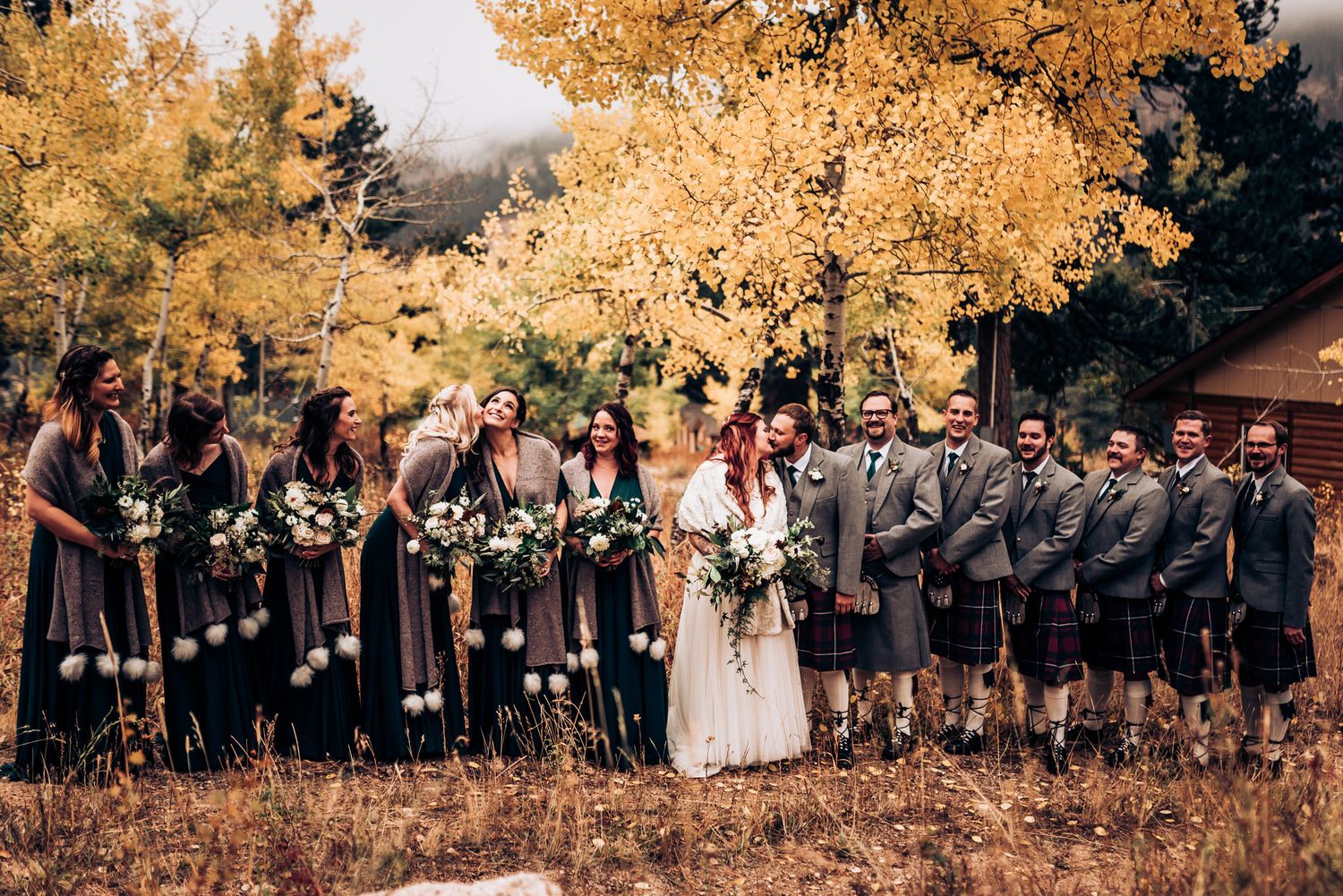 Large wedding party in kilts and formal attire stands together under golden autumn trees.