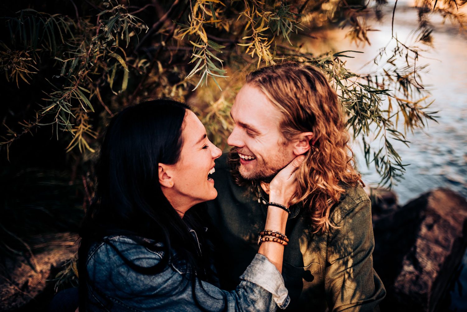 Couple shares intimate laugh together under tree branches by waterfront.