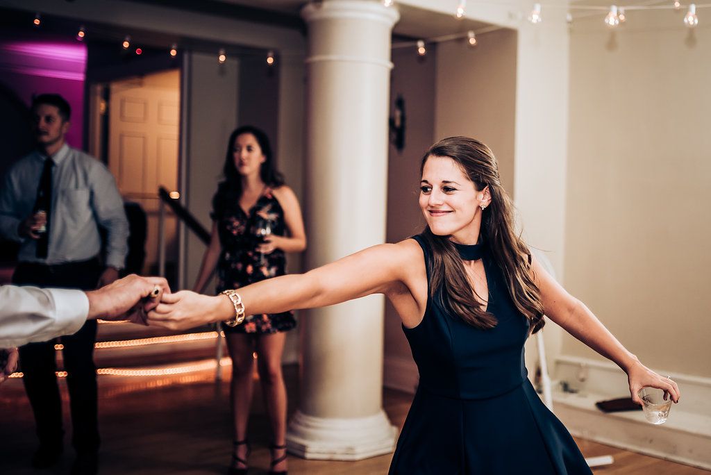 People dancing and celebrating at an indoor event venue with columns and string lights.
