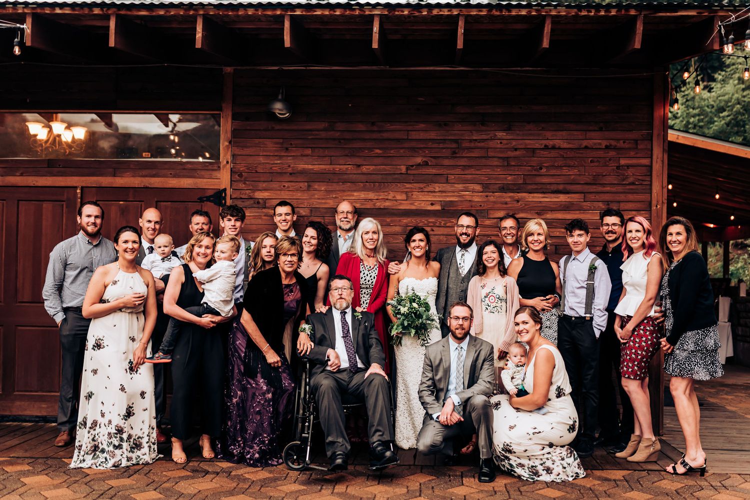 Large wedding party group photo in front of rustic wooden venue with string lights overhead.