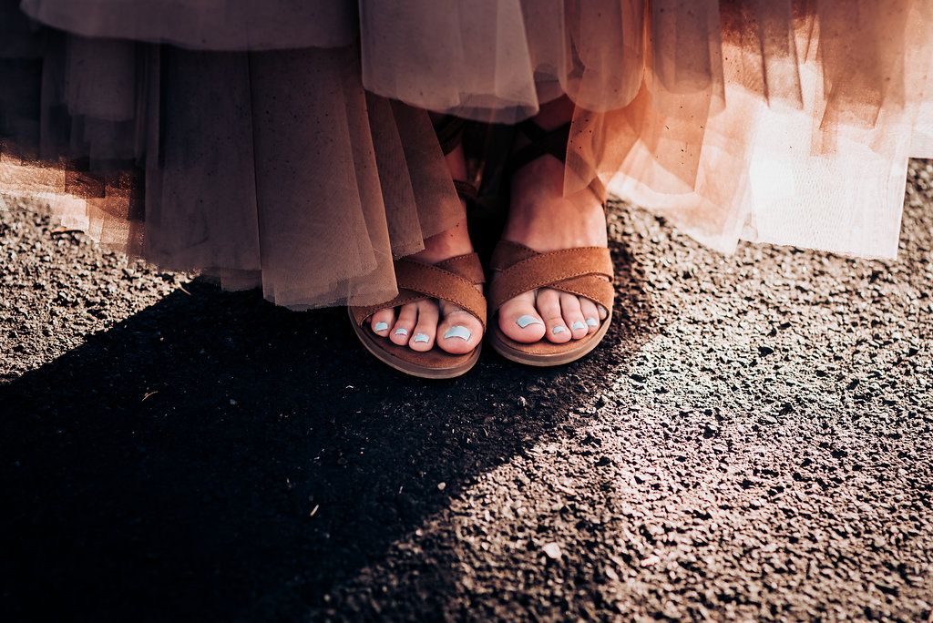 Close-up of sandaled feet beneath flowing dress fabric on a sunlit rocky surface.