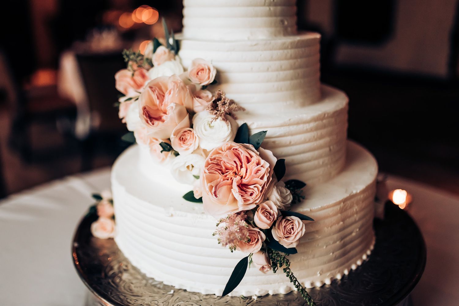 Four-tier white wedding cake adorned with delicate peach roses and dark green foliage.