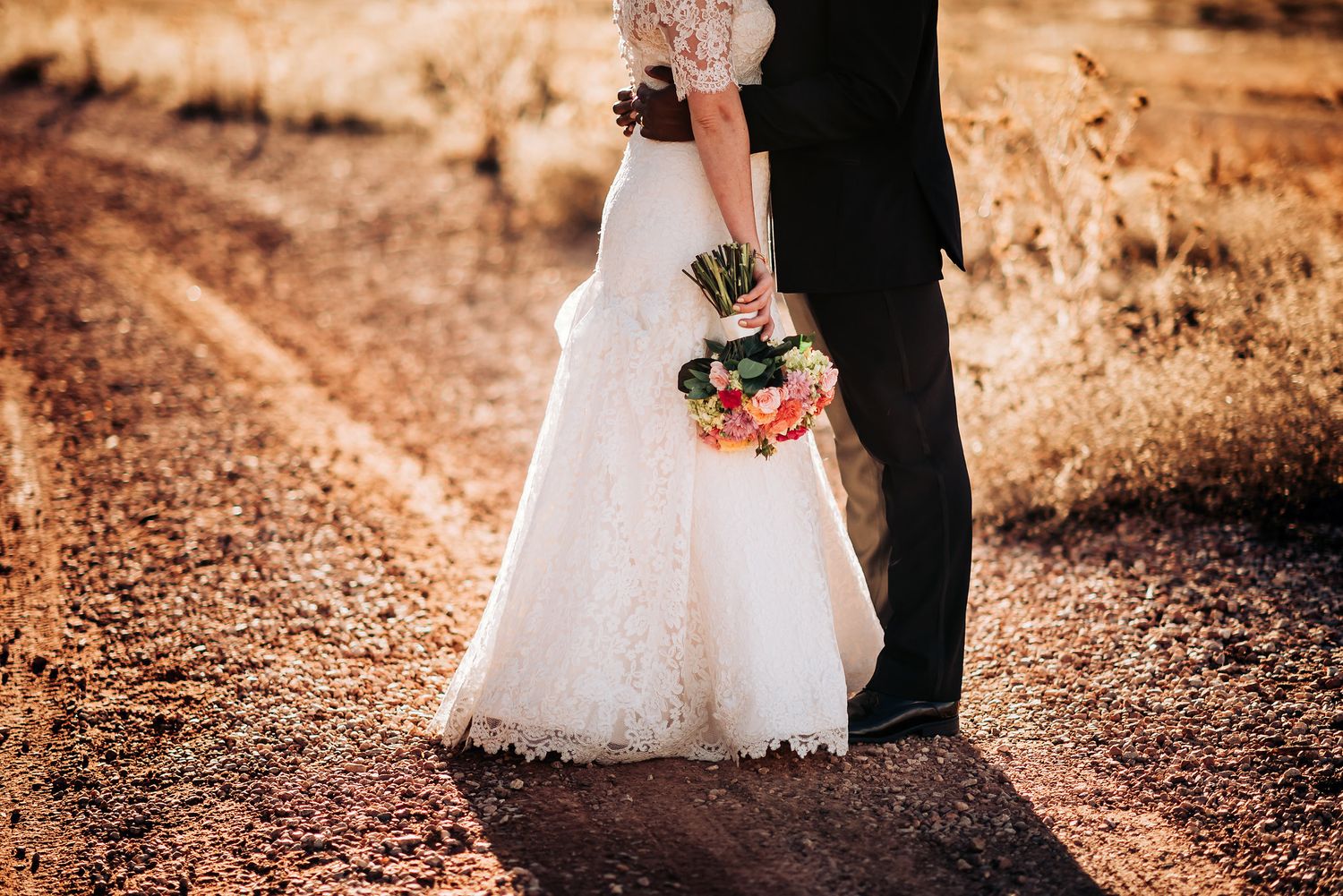 Close-up of bride's lace dress and bouquet while standing with groom on dirt path.