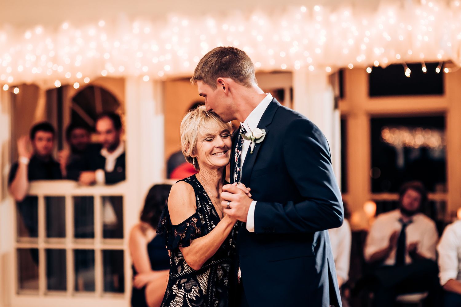 A couple enjoys a romantic dance moment at a wedding reception with string lights overhead.